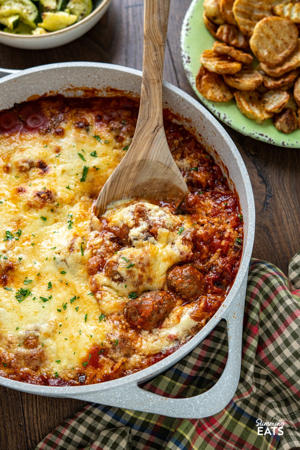 Indulge in the tantalizing flavors of the Mediterranean with this delicious creation of Moussaka Meatballs - tender seasoned lamb meatballs in a rich tomato sauce topped with a light creamy Bechamel sauce. Get ready to embark on a culinary journey that will transport your taste buds straight to the sun-soaked shores of Greece! Greek Moussaka Meatballs in a non stick ceramic casserole dish with potato and courgette sides