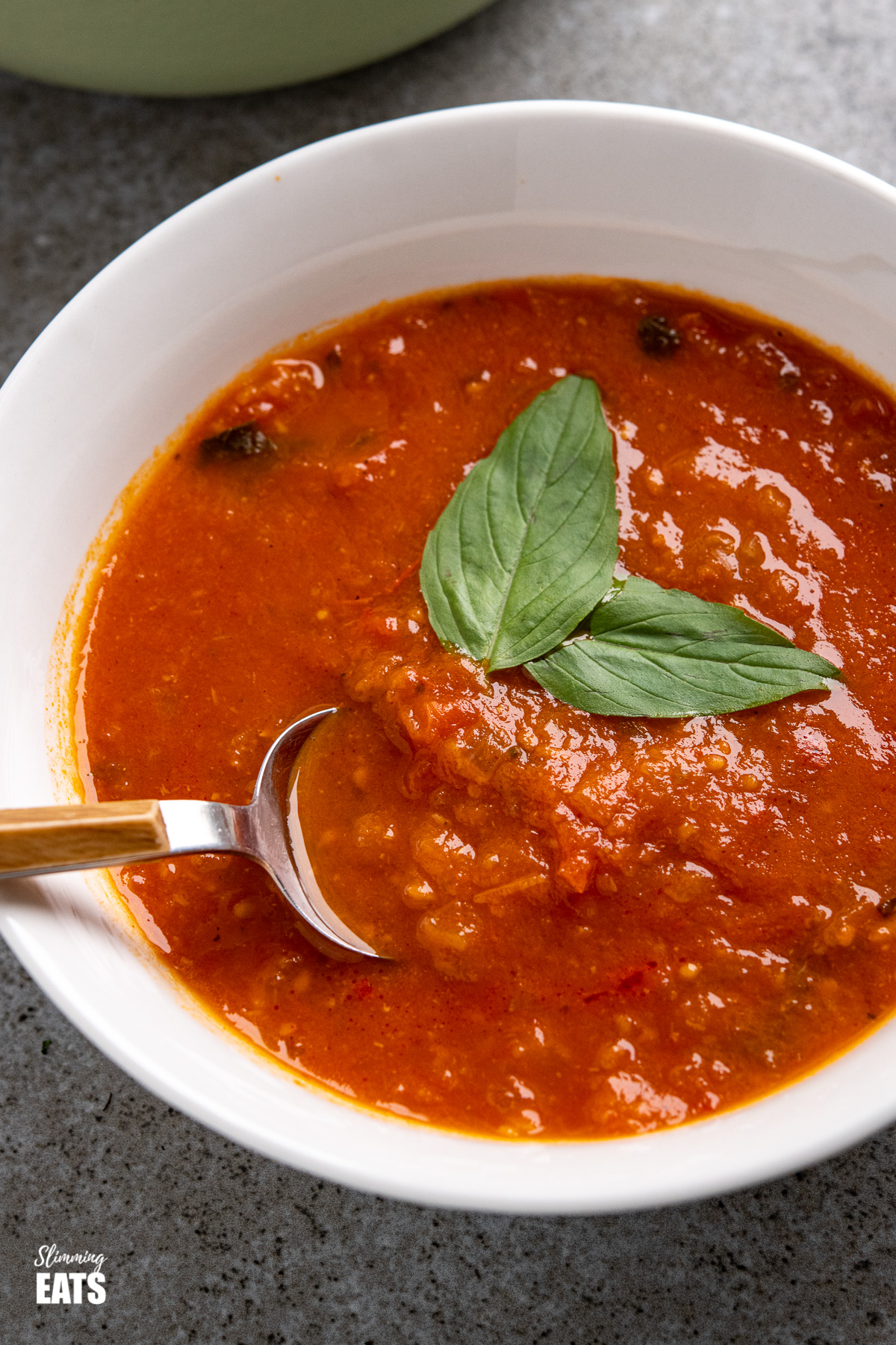 close up of white bowl of Fresh Tomato and Basil Soup with spoon with wooden style handle, garnished with fresh basil leaves
