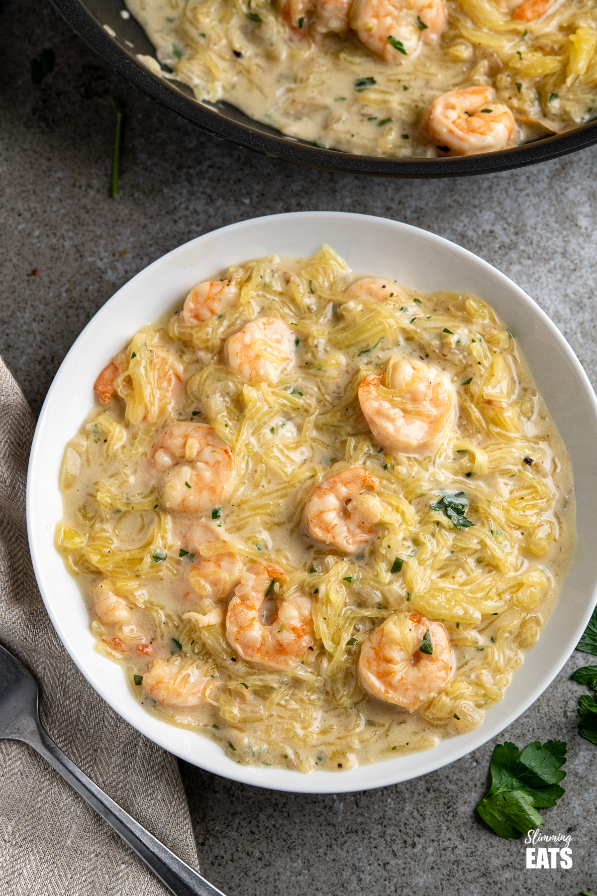 Shrimp Spaghetti Squash Alfredo in a white bowl with scattered parsley and frying pan above, plus fork placed to the left.