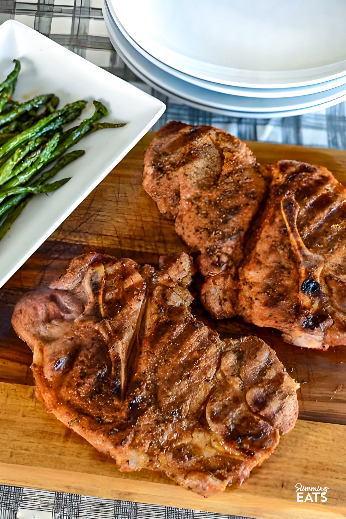 Grilled BBQ pork chops served on a wooden board, with white plates in the background showcasing a side of asparagus.