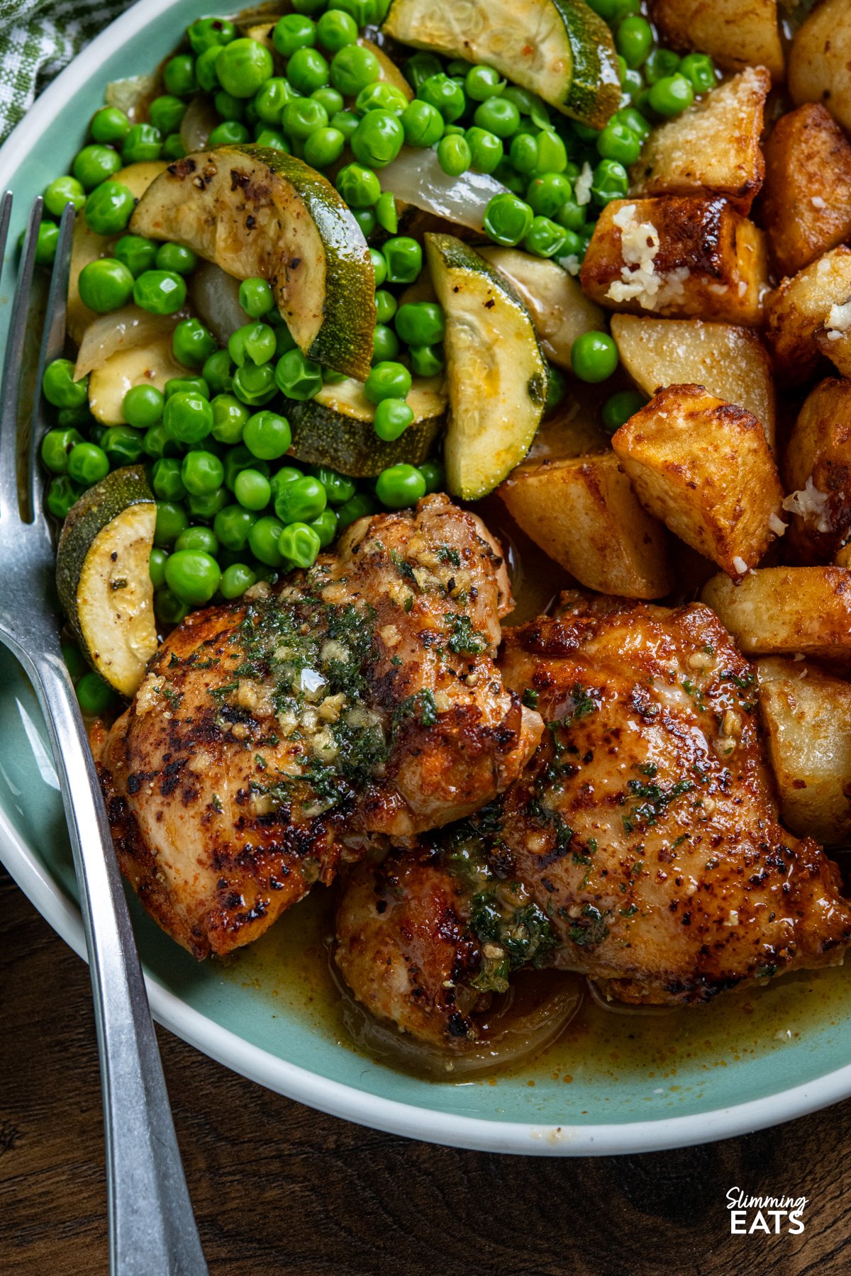 This Lemon Garlic Chicken and Zucchini Bake is an easy, delicious meal. Juicy chicken is nestled in a zesty lemon and garlic butter sauce with tender seasoned zucchini. close up of Garlic Lemon Chicken and Zucchini Bake served on a turquoise plate alongside golden parmesan potatoes and garden peas, fork placed on left hand side of plate.