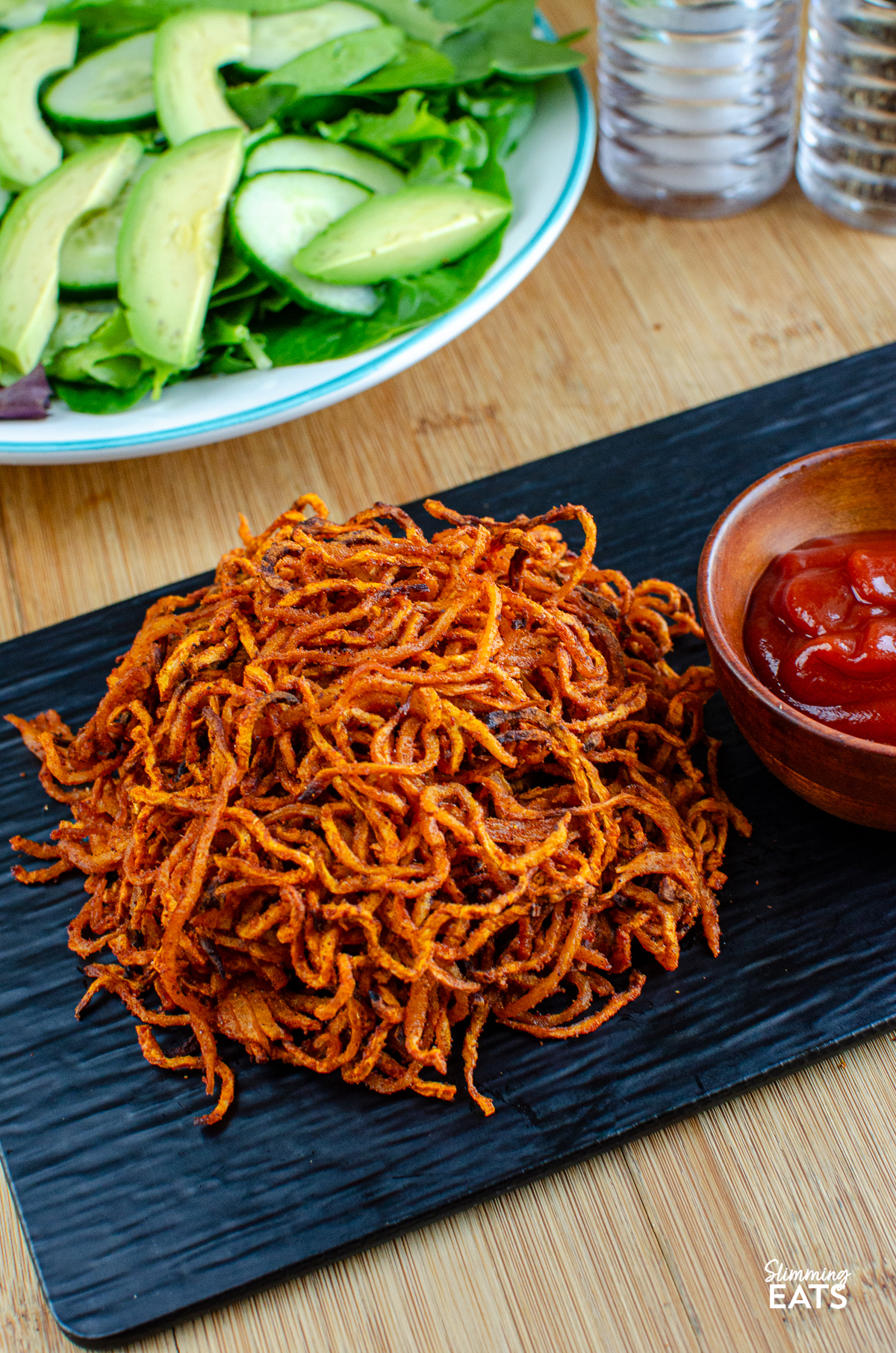 spiralized seasoned swede fries on a black slate tray with a wooden bowl with ketchup, bowl of mixed salad and salt and pepper in background