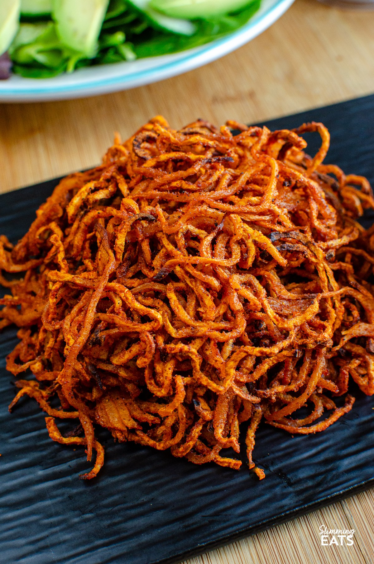 close up spiralized seasoned swede fries on a black slate tray, bowl of mixed salad and salt and pepper in background