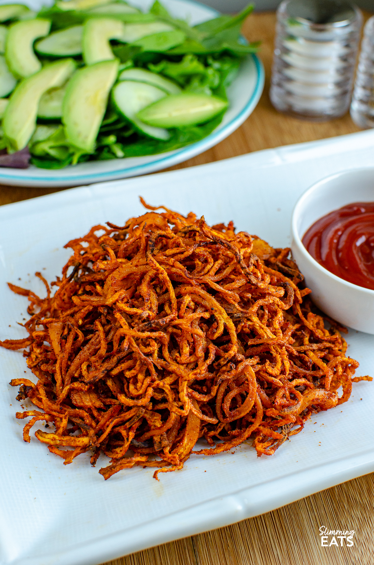 spiralized seasoned swede fries on a white rectangle plate with white bowl with ketchup, bowl of mixed salad and salt and pepper in background