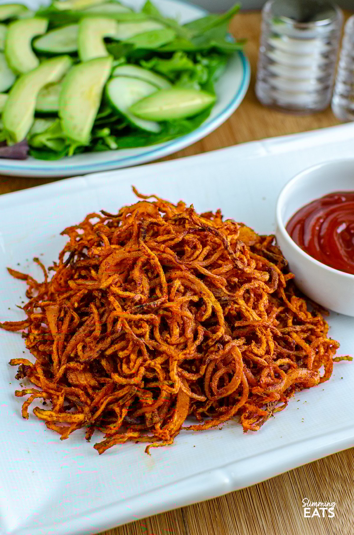 Spiralized Seasoned Swede Fries - these delightful spirals of swede offer a nutritious twist to traditional fries, ideal for those seeking a lower-carb alternative to accompany their high-protein main course. spiralized seasoned swede fries on a white rectangle plate with white bowl with ketchup, bowl of mixed salad and salt and pepper in background