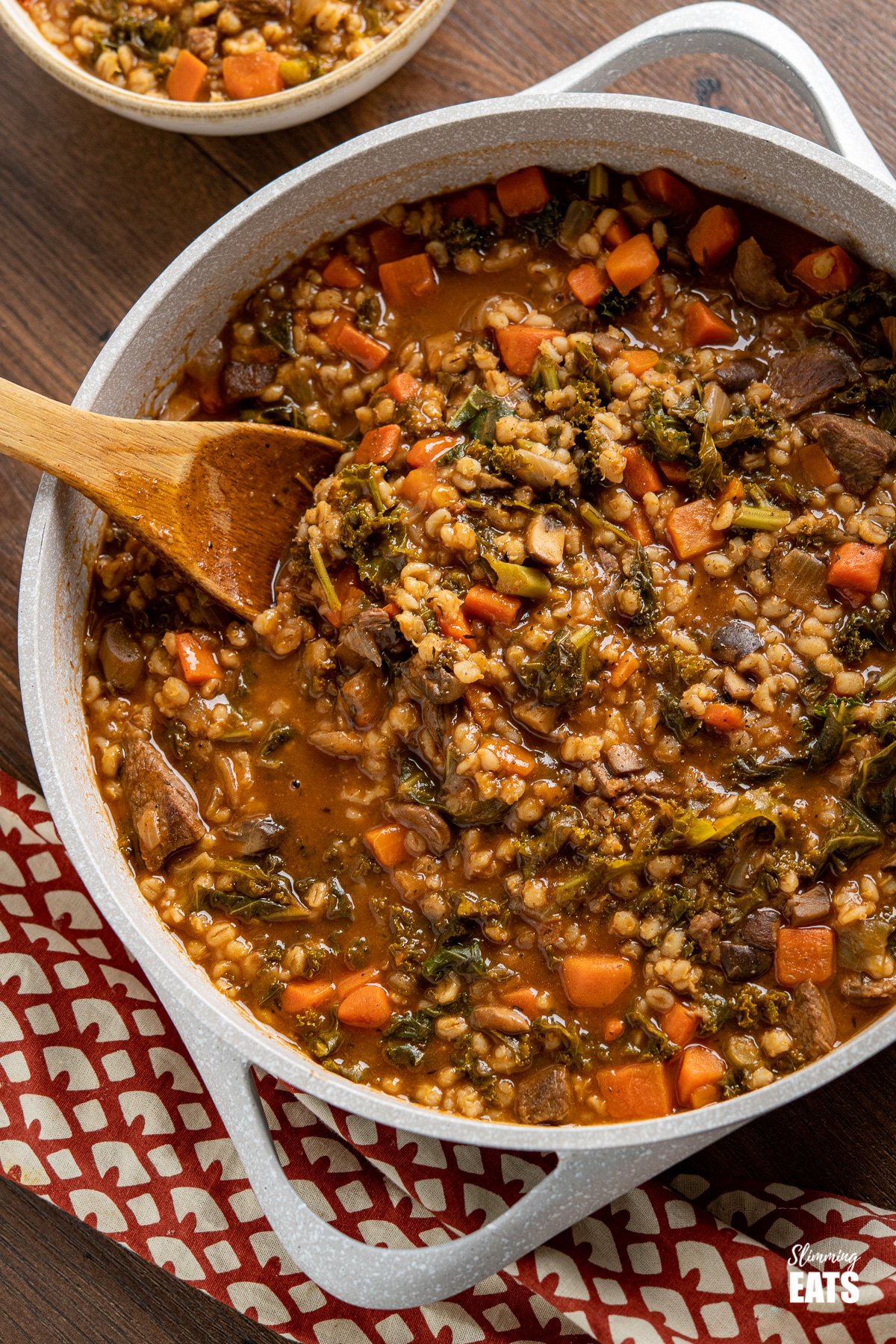 Beef and barley stew in pot with wooden spoon, bowl of stew in background