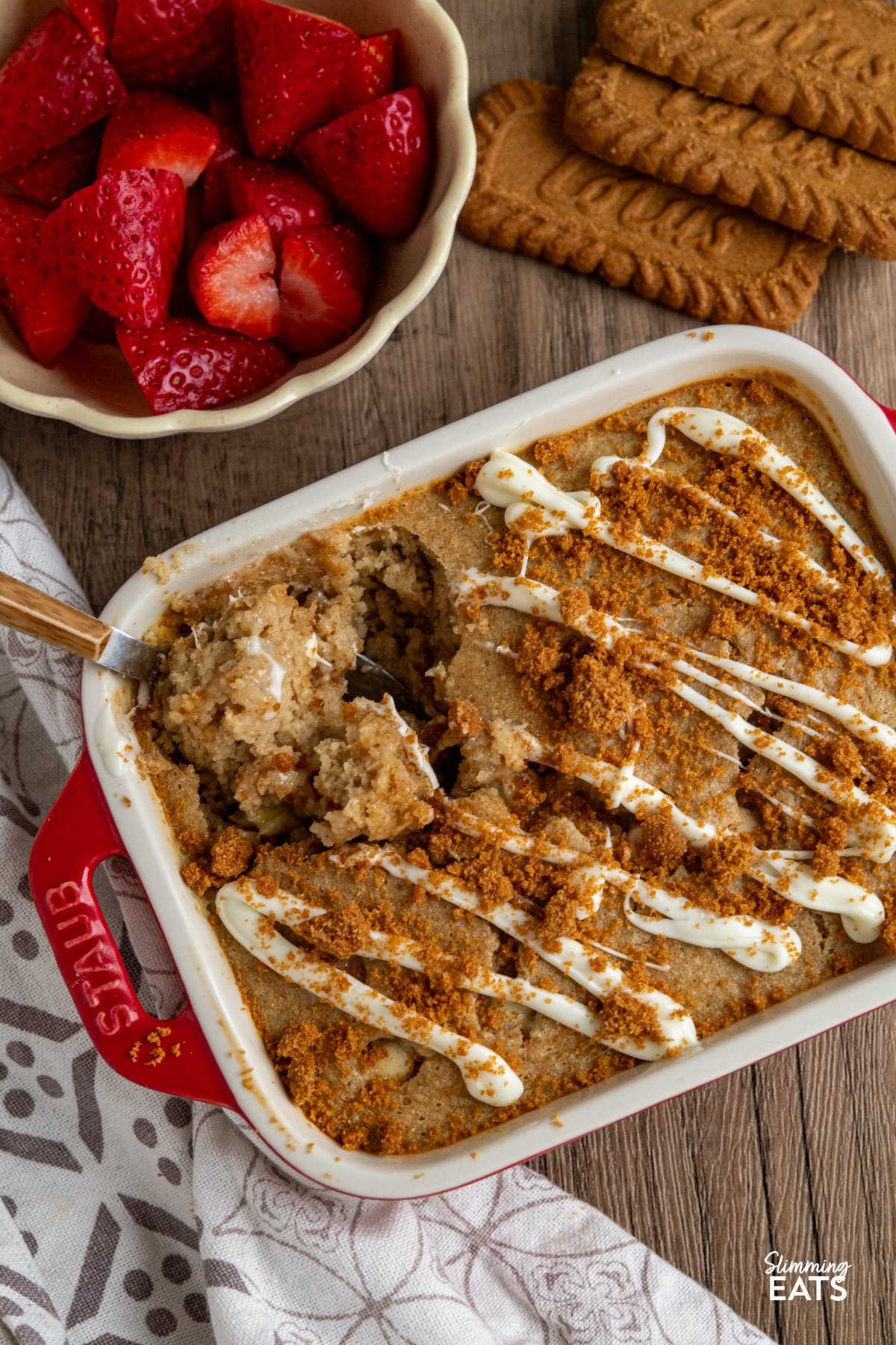 spoon in White Chocolate Chip Oatmeal Cake with Biscoff Cookie Crumb in red mini staub baking dish  surrounded by lotus biscuits and bowl of strawberries. 