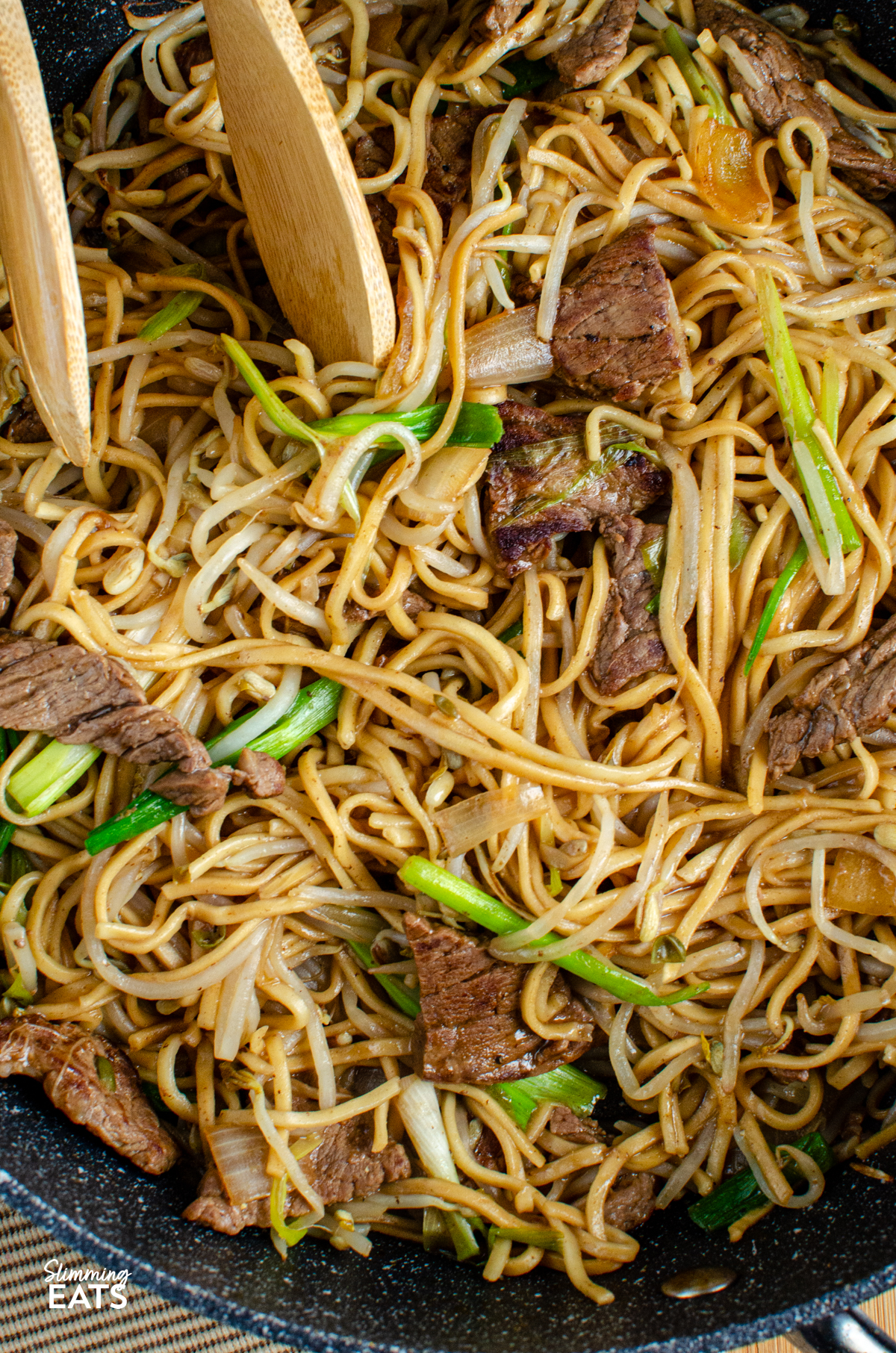 close up of Beef Chow Mein in a non stick frying pan with wooden tongs
