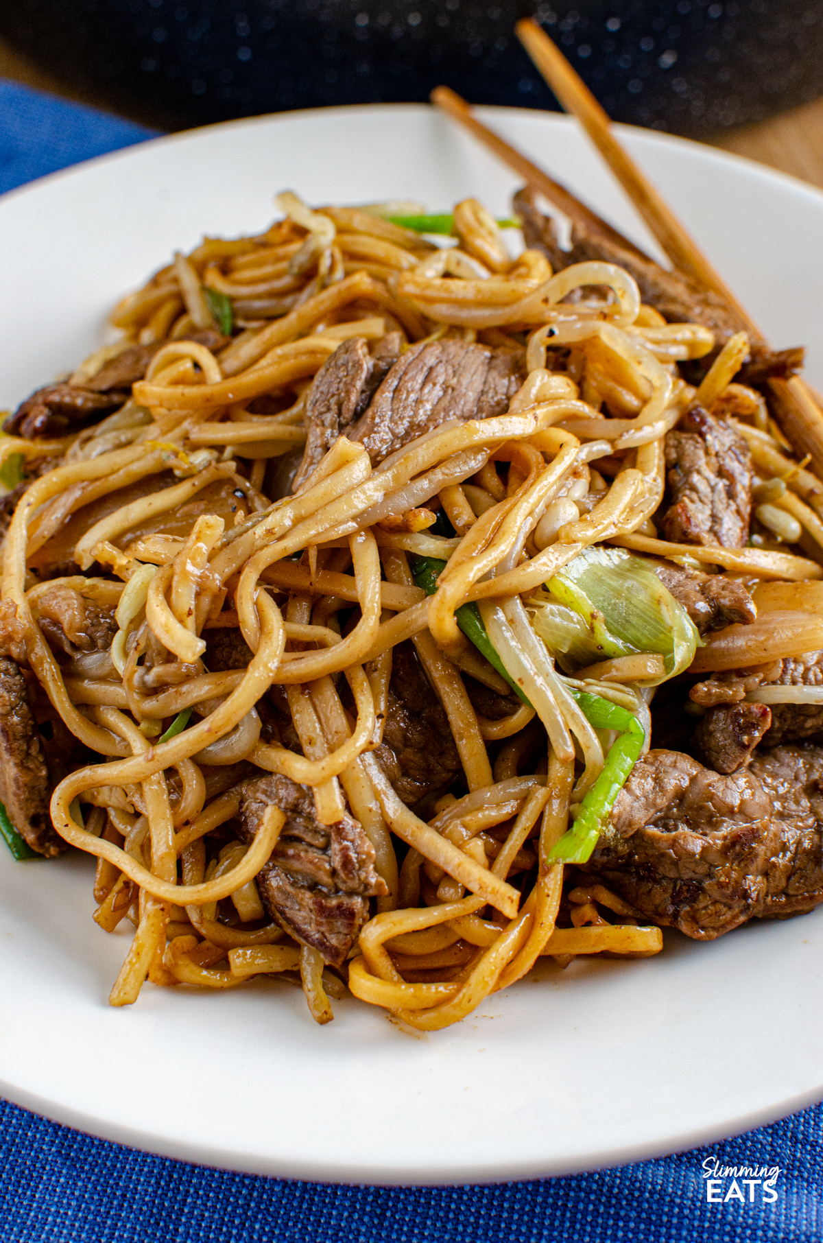 close up of serving of Beef Chow Mein  on a white plate with chopsticks, placed on a blue napkin