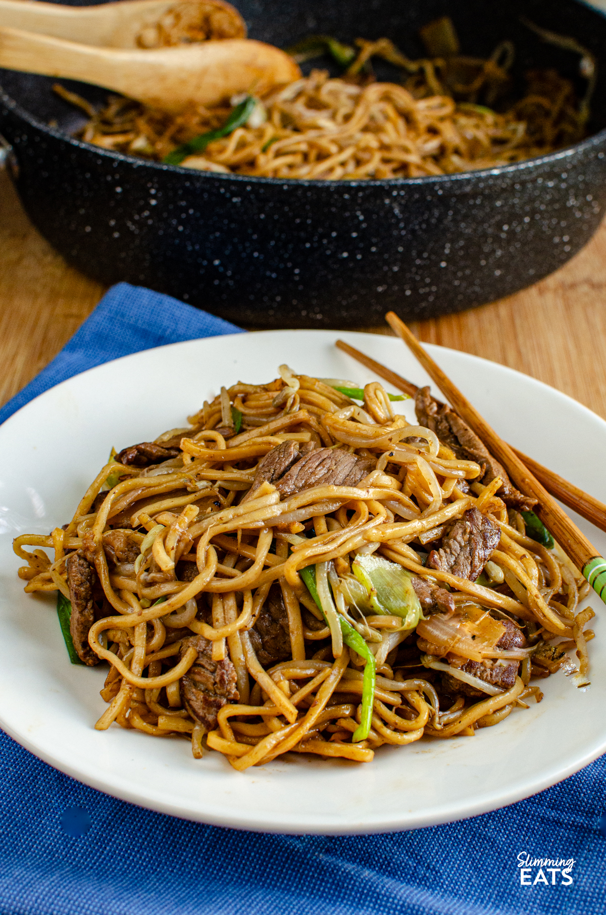 serving of Beef Chow Mein  on a white plate with chopsticks, placed on a blue napkin, frying pan visible in background