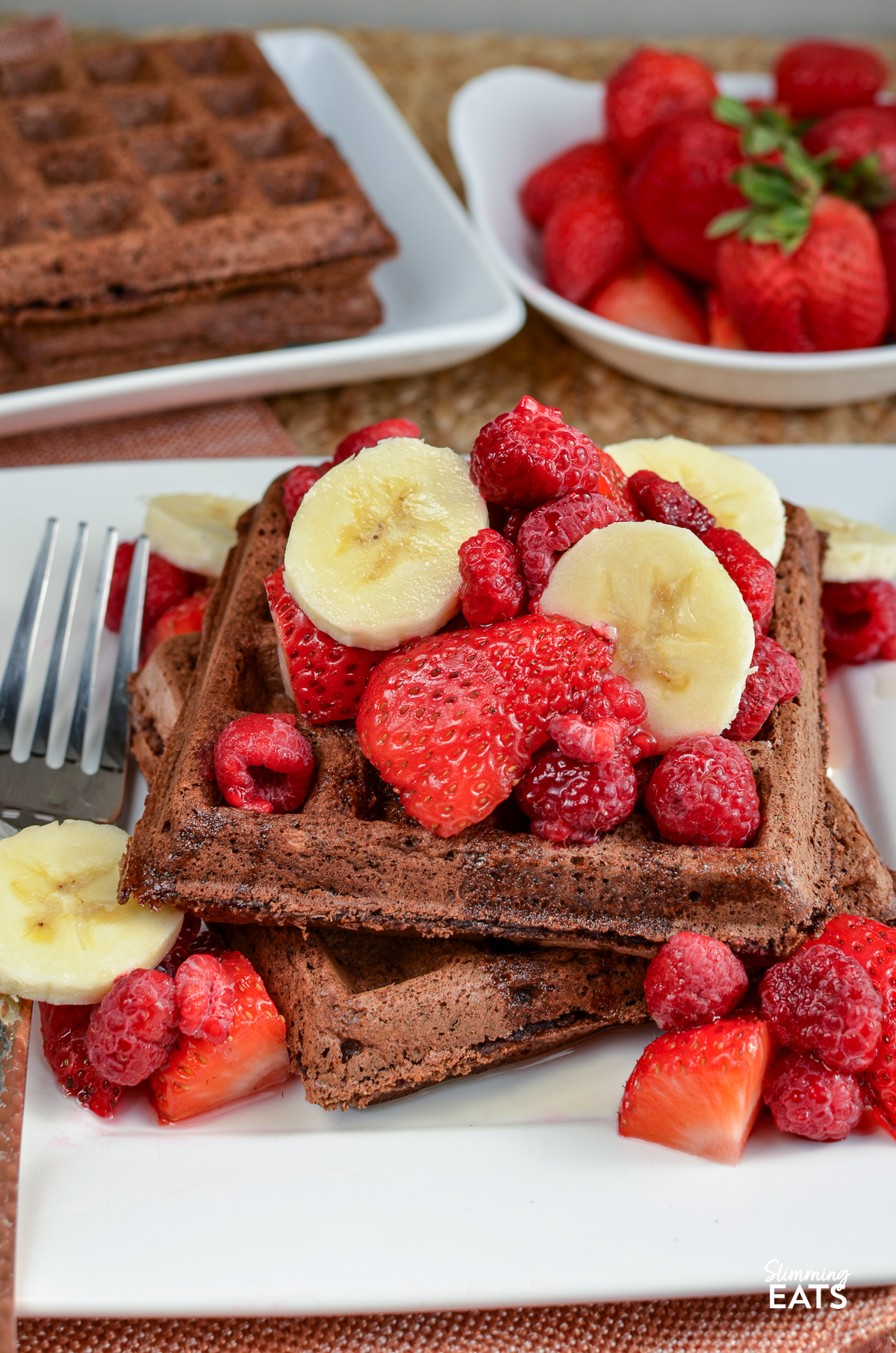 Chocolate oat banana waffles served on a white square plate adorned with fresh banana and strawberry slices, accompanied by a decadent drizzle of maple syrup. Additional toppings are visible in the background