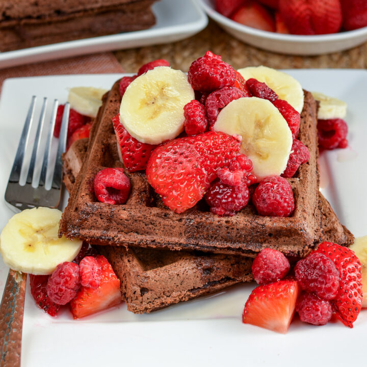 Chocolate oat banana waffles served on a white square plate adorned with fresh banana and strawberry slices, accompanied by a decadent drizzle of maple syrup. Additional toppings are visible in the background