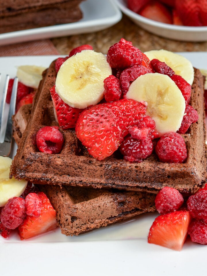 Chocolate oat banana waffles served on a white square plate adorned with fresh banana and strawberry slices, accompanied by a decadent drizzle of maple syrup. Additional toppings are visible in the background