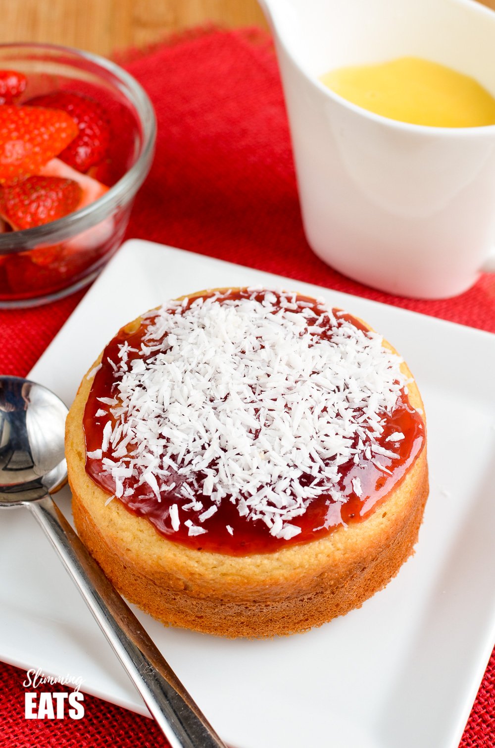 Jam and Coconut Sponge Cake - a delicious journey back to childhood with this oat based coconut sponge, topped with sweet jam and shredded coconut. A perfect filling start to your day. Gluten Free, Vegetarian, Slimming and Weight Watchers friendly jam and coconut sponge cake on a white plate and spoon with strawberries and jug of custard