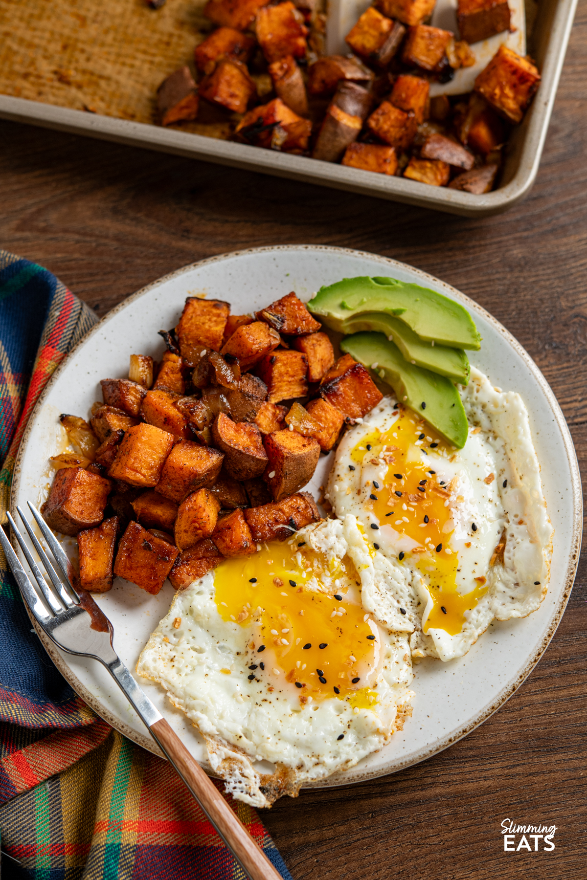 roasted skin on seasoned sweet potato cubes with onion on a white plate with 2 fried eggs and avocado slices