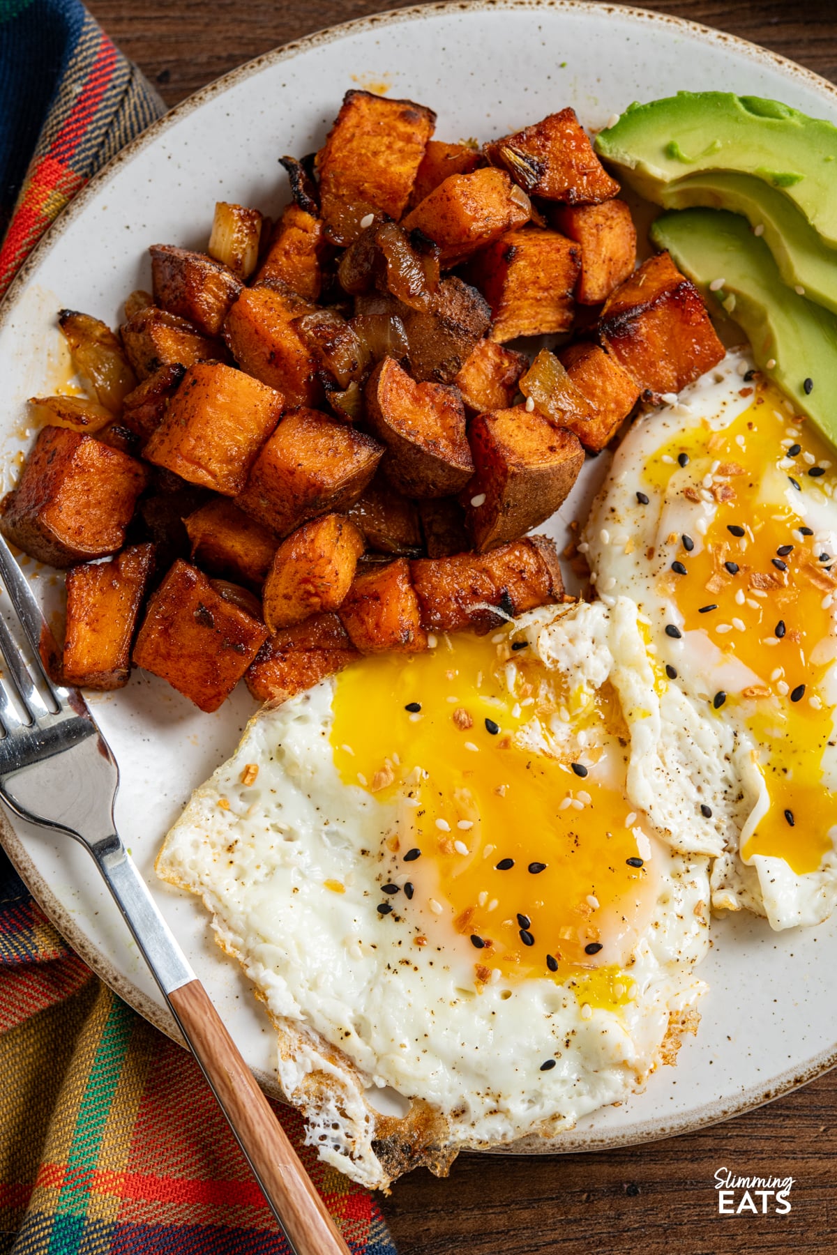 close up of roasted skin on seasoned sweet potato cubes with onion on a white plate with 2 fried eggs and avocado slices