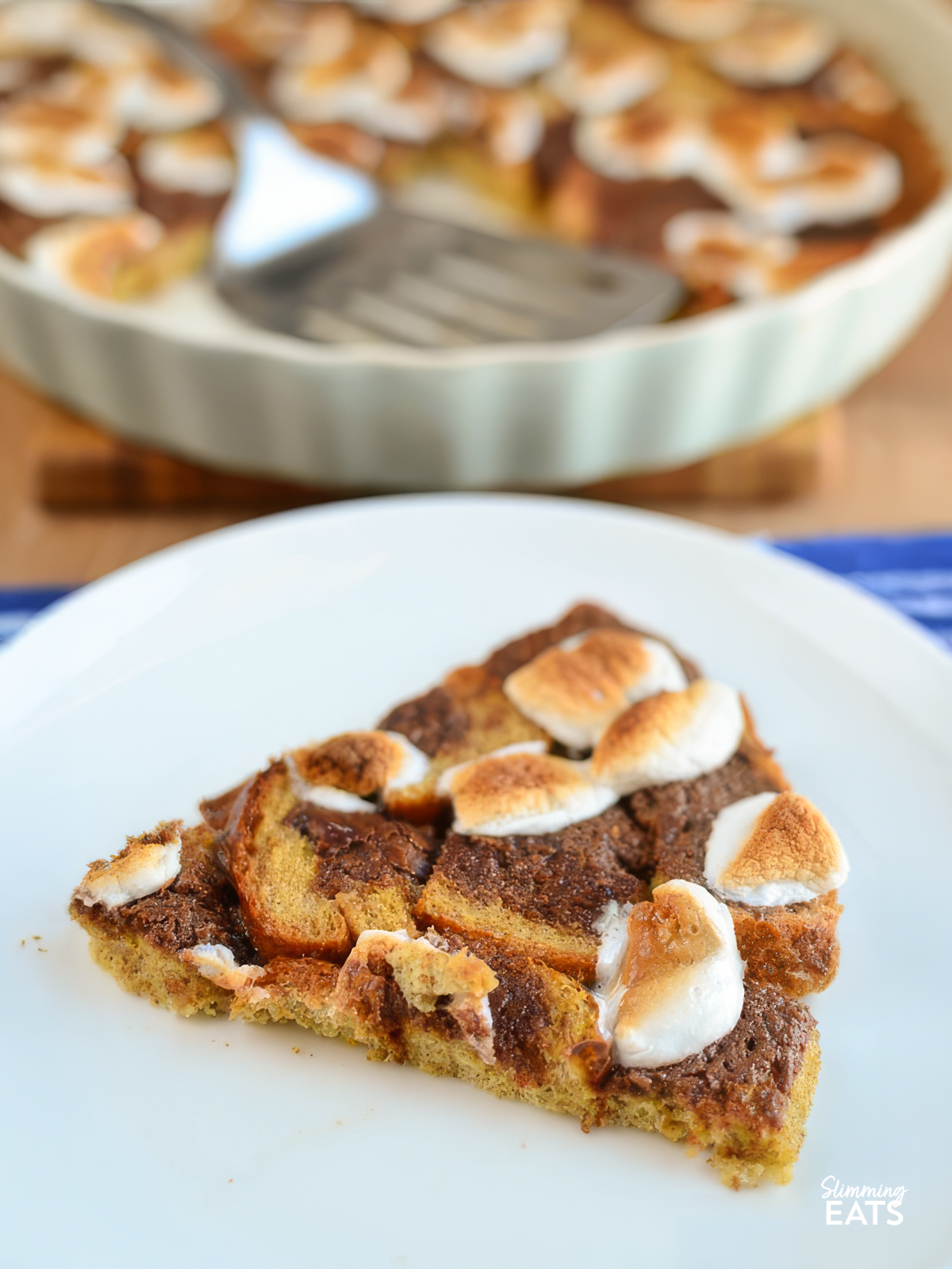 smores bread pudding portion on a white plate with the remainder in a ceramic pie dish in background