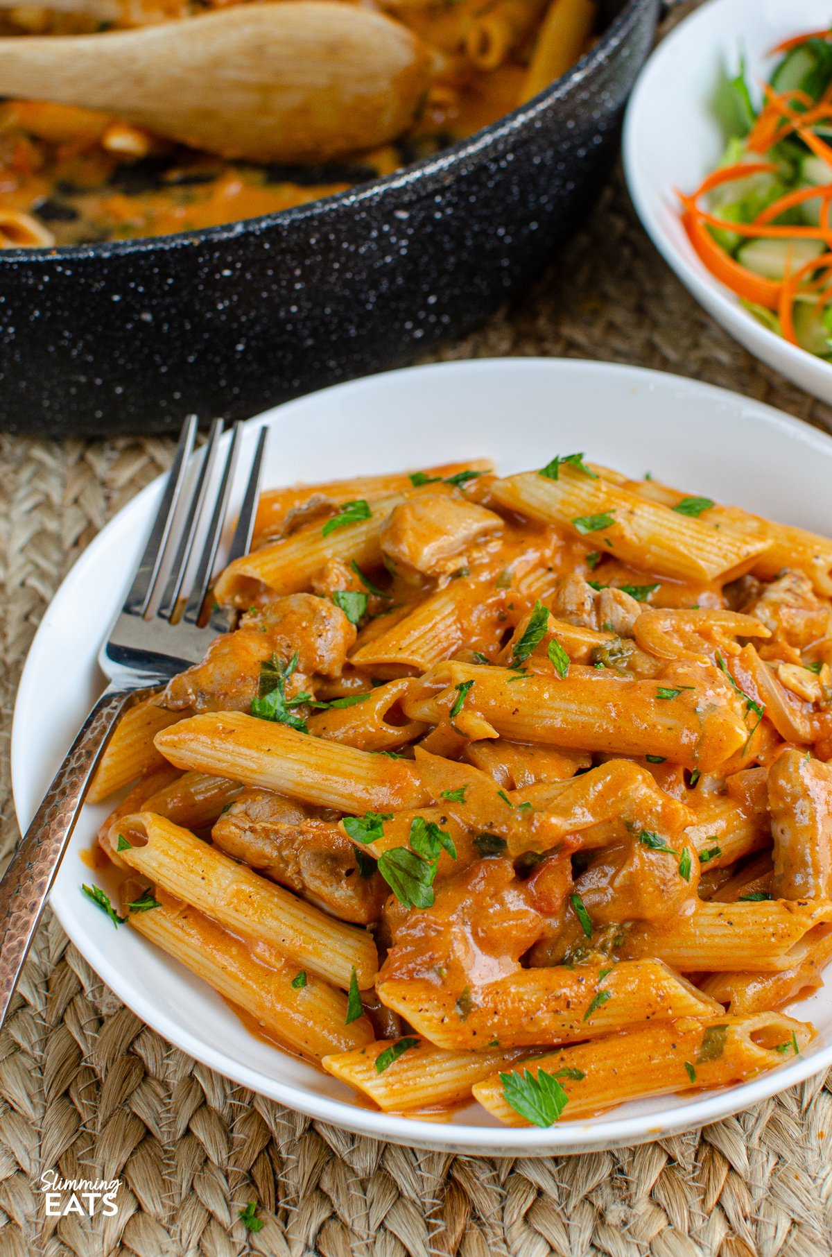 creamy chicken and tomatoes mixed with pasta in a white bowl, frying pan and bowl of salad in background