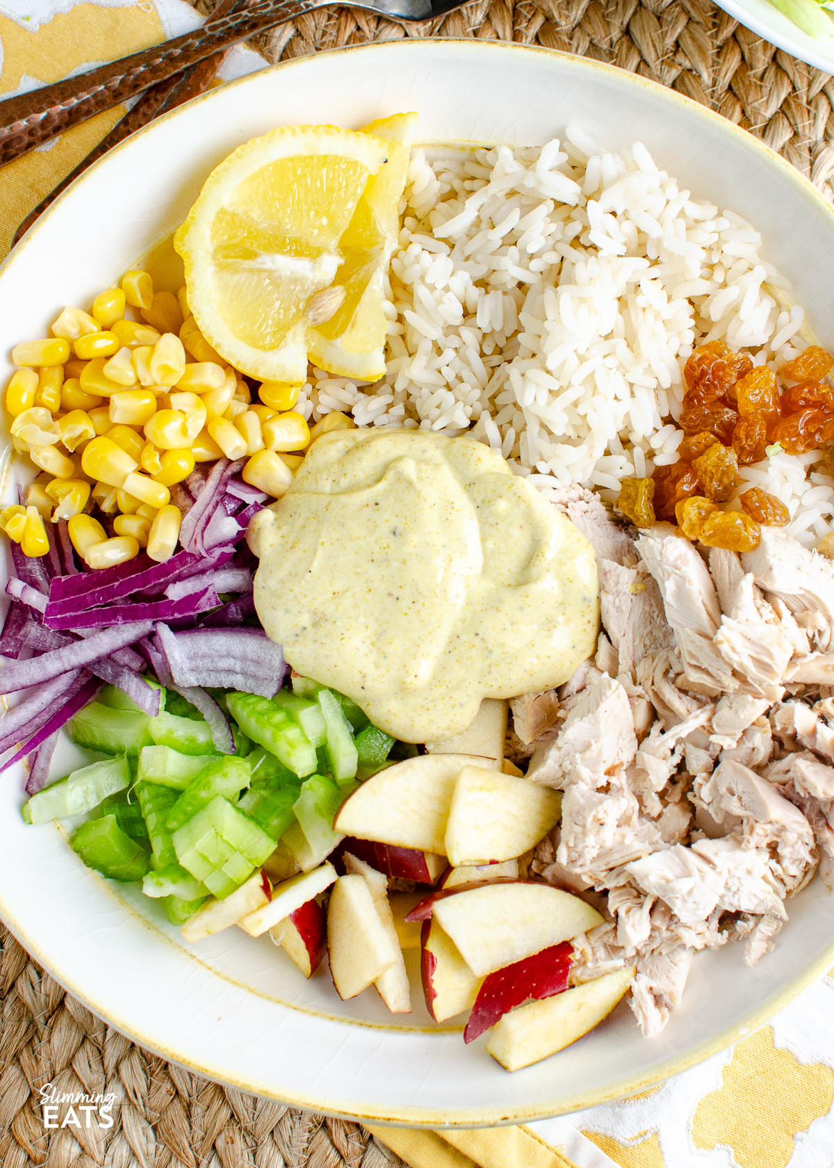 Ingredients for curried chicken rice salad bowl arranged side by side, unmixed, in a bowl. Dressing spooned in the middle, with forks placed on a  mat above