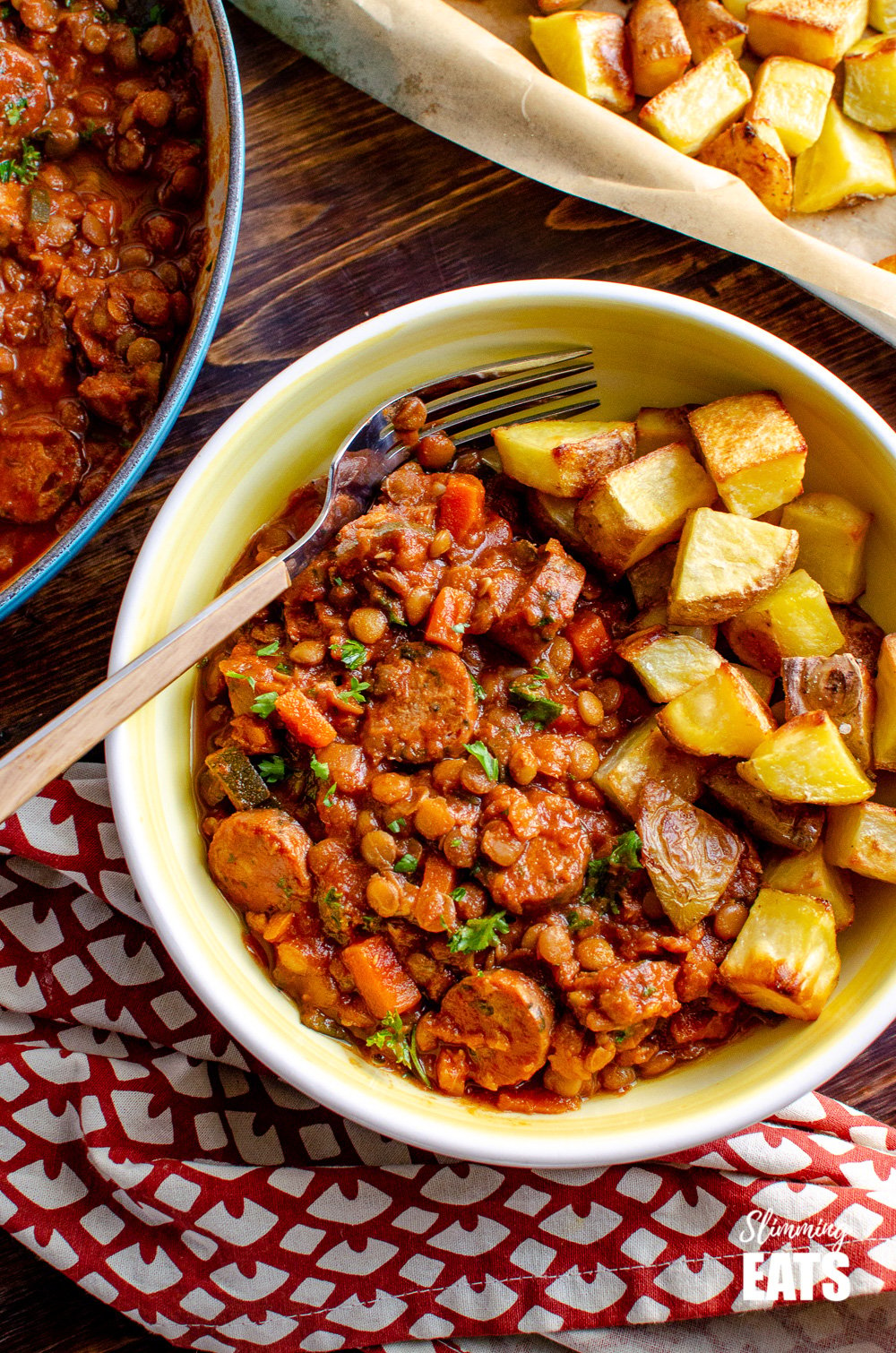 over top view of One Pot Sausage and Lentil Casserole in white and yellow bowl with roast potatoes and wooden handled fork