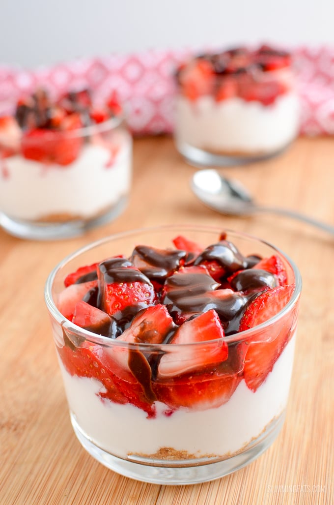 Strawberry Topped Greek Yoghurt Style Cheesecakes - Enjoy one of these delicious Strawberry Cheesecakes that are super quick to make and a delicious treat. Slimming Eats and Weight Watchers friendly 3 strawberry Greek yoghurt cheesecake in glass dishes place on a wooden board with spoon