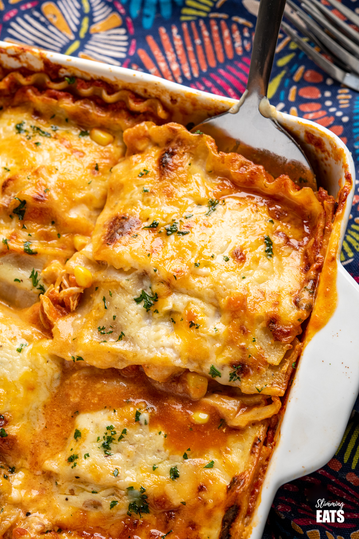 spatula scooping up chicken and sweetcorn lasagne in a white oven proof dish