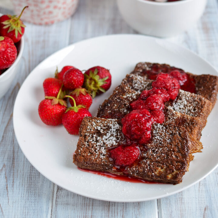Chocolate French toast with strawberry syrup and fresh strawberries served on a white plate.