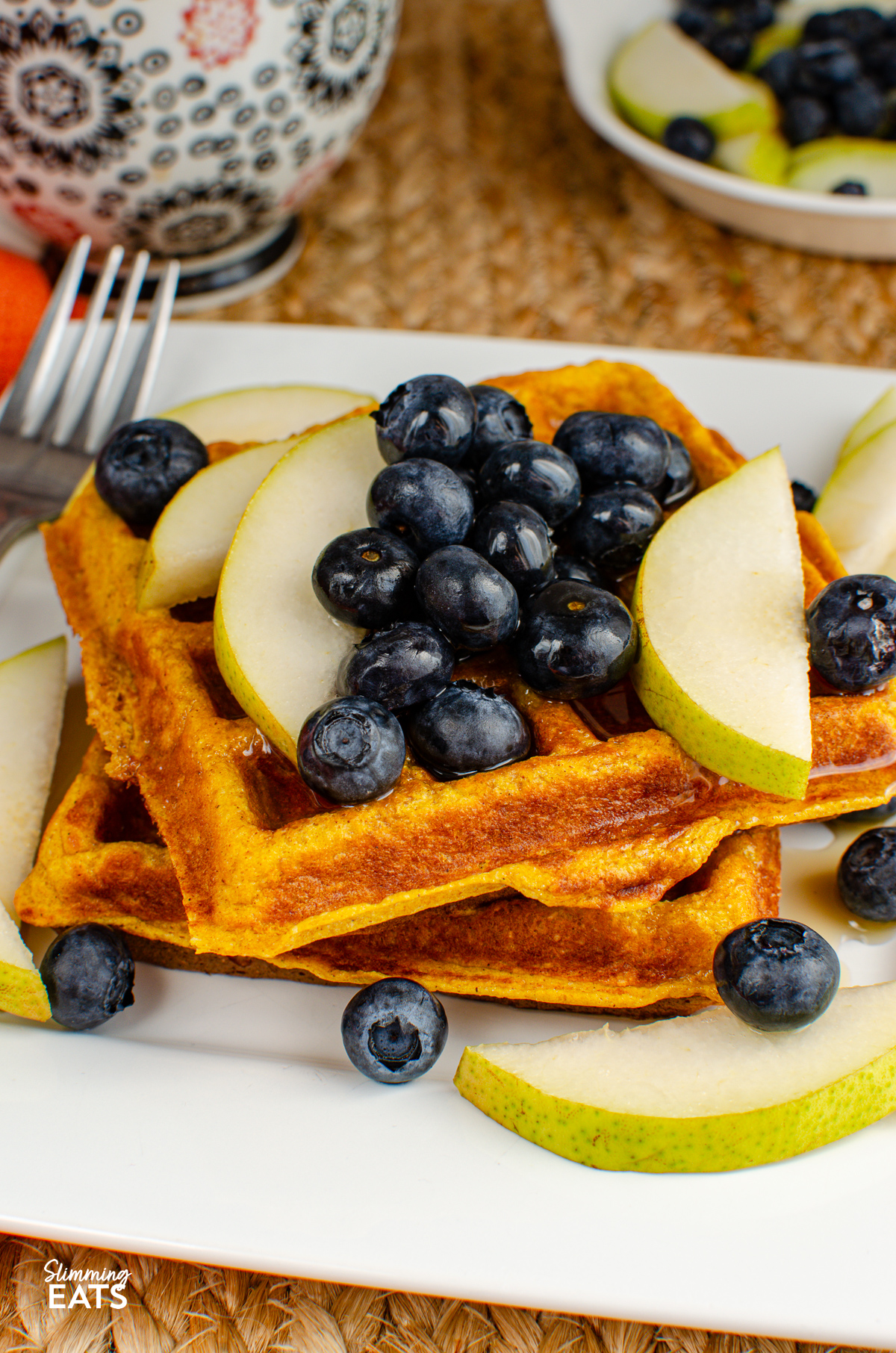 Delicious homemade oat pumpkin waffles served on a pristine white plate, adorned with fresh blueberries and slices of ripe pear, perfectly complemented by a delicate drizzle of golden maple syrup.