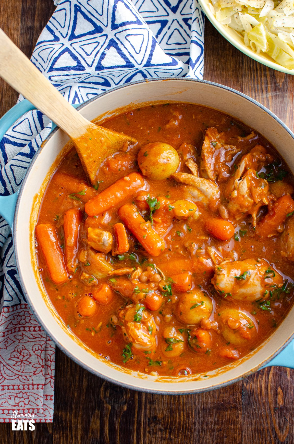 Chicken and Baked Bean Casserole - fall off the bone meat, tender pieces of chicken with delicious vegetables of carrots and new potatoes in a rich baked bean and tomato sauce. #chicken #potatoes #beans #casserole #instantpot #slowcooker #slimmingeats #weightwatchers over the top view of chicken and baked bean casserole in cast iron pot with cabbage in a bowl above