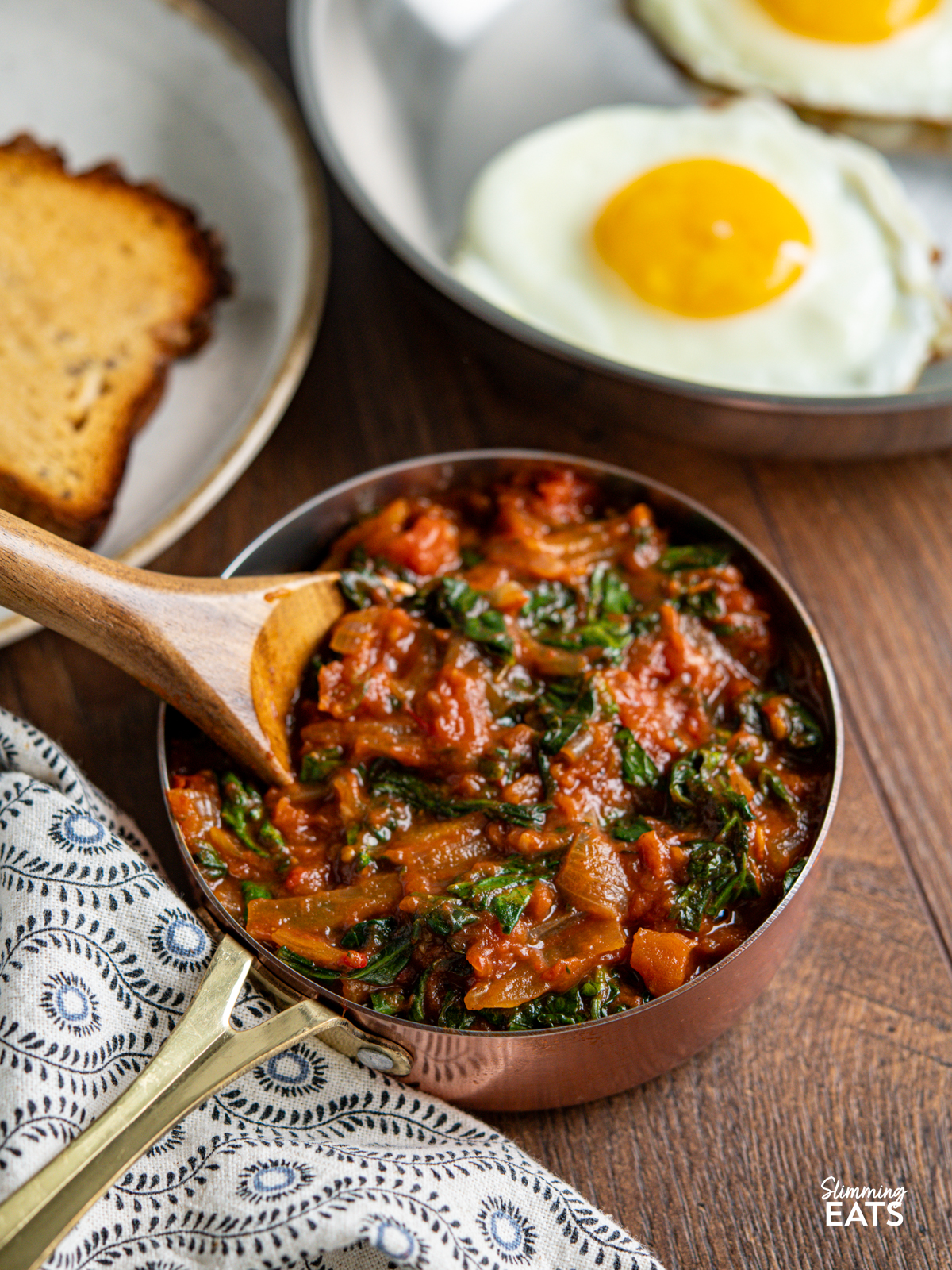 balsamic tomatoes and spinach in a mini copper saucepan with wooden spoon, eggs in pan and toast on plate in background