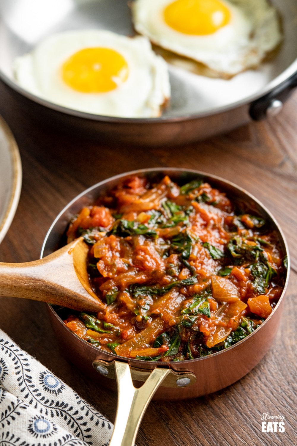 balsamic tomatoes with spinach in a copper mini saucepan with frying pan of eggs in background