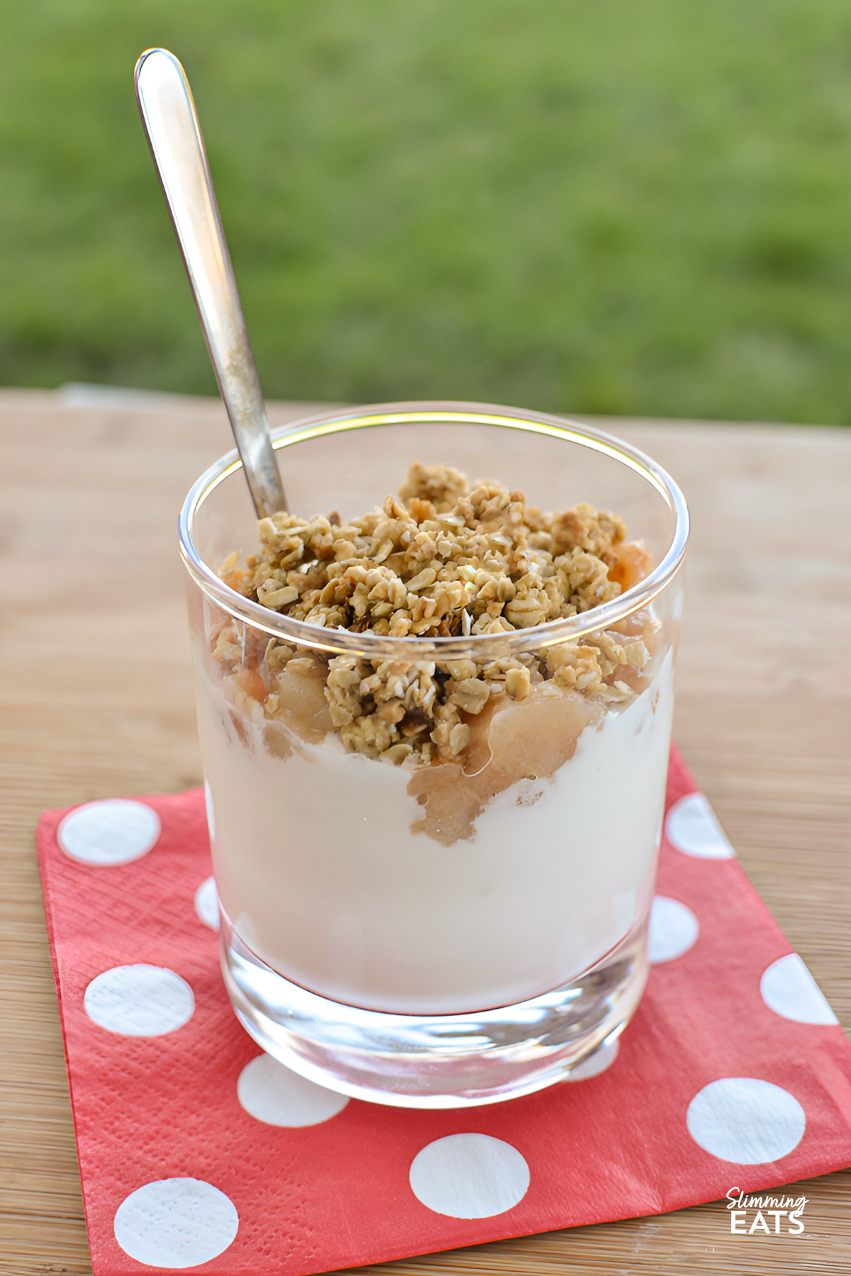A Small glass filled with layers of pear crumble and yogurt parfaits, accompanied by a spoon, resting on a pale red napkin adorned with white polka dots