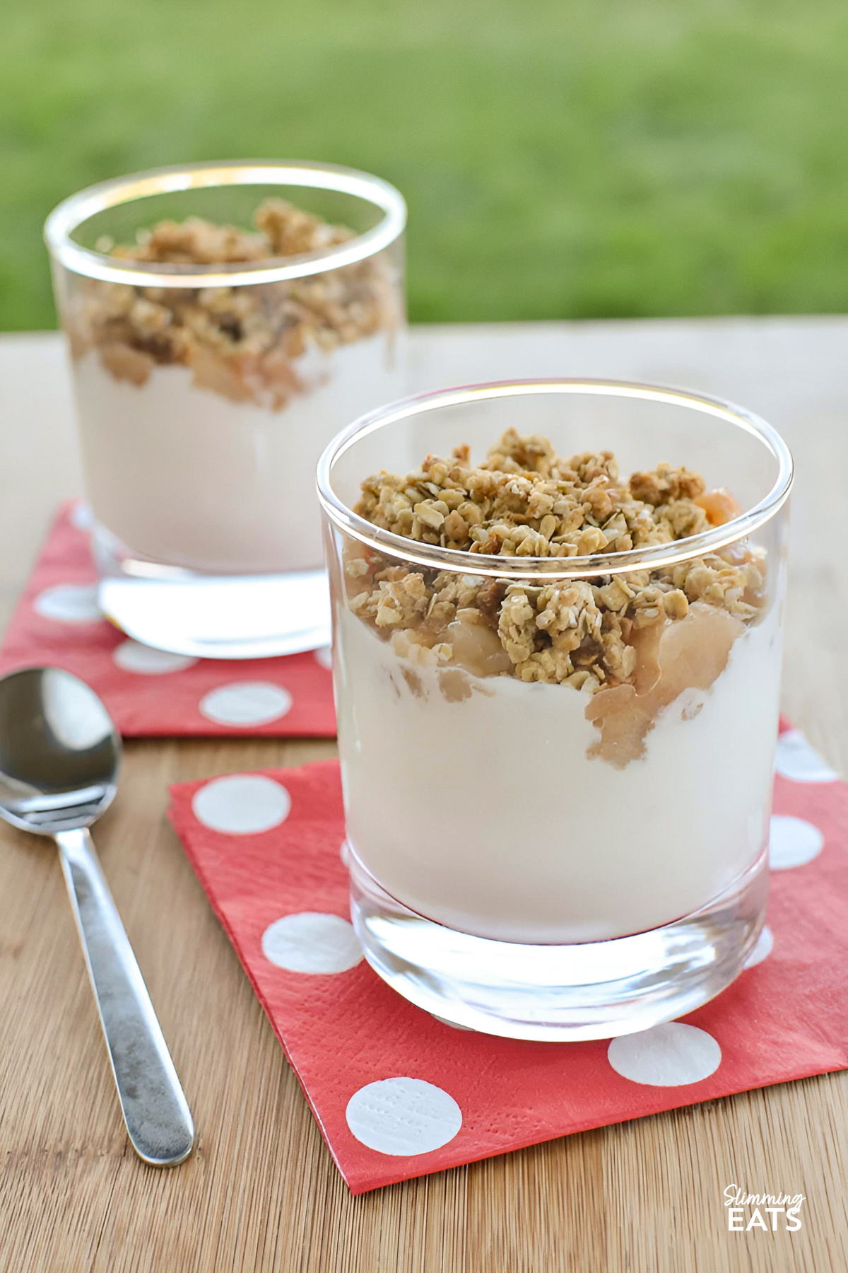 Small glasses filled with layers of pear crumble and yogurt parfaits, accompanied by a spoon, resting on a pale red napkin adorned with white polka dots