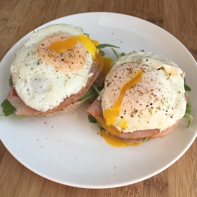 cheats poached eggs over ham and baby greens on whole wheat English muffins on a white plate