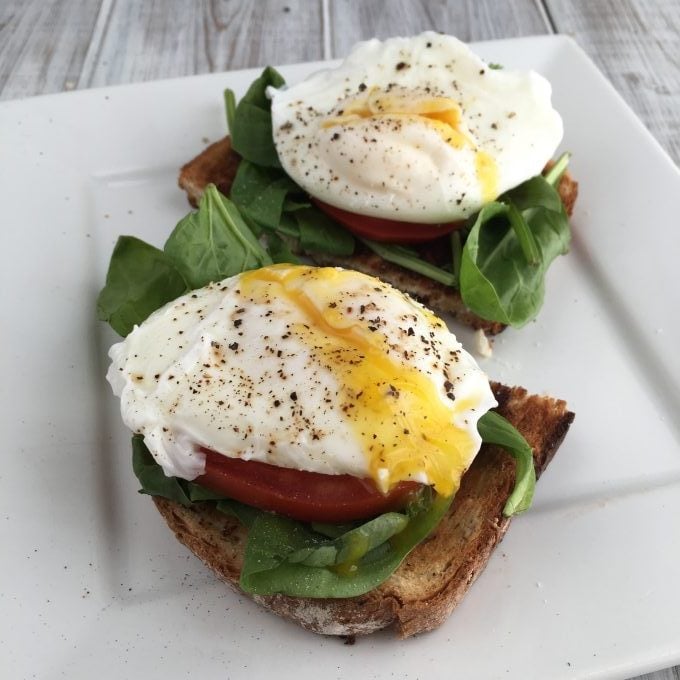 cheats poached eggs over tomato slices, baby greens on whole wheat toast on a white plate
