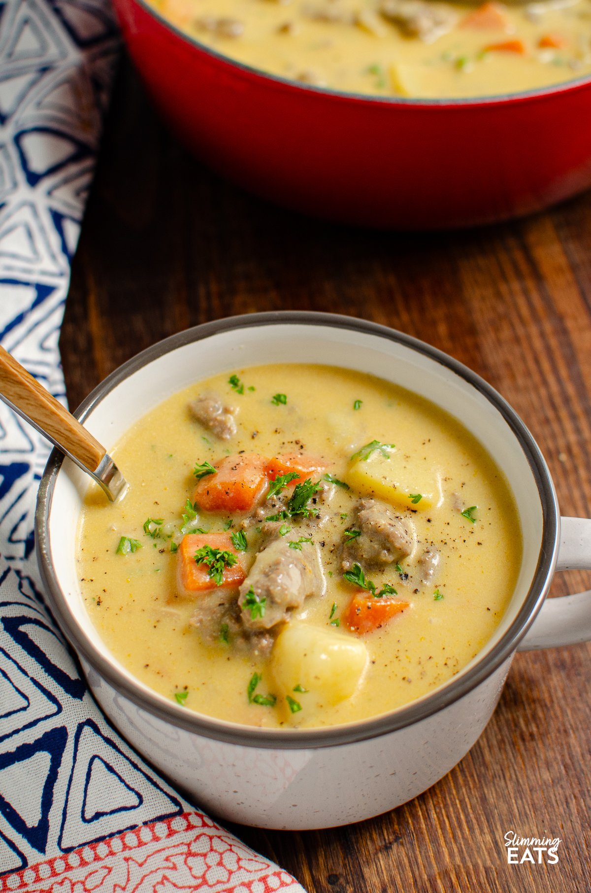 A close-up of a grey-brown ceramic mug filled with Cheeseburger Soup, topped with a spoon. In the background, a rustic pot of soup simmers on a stove.