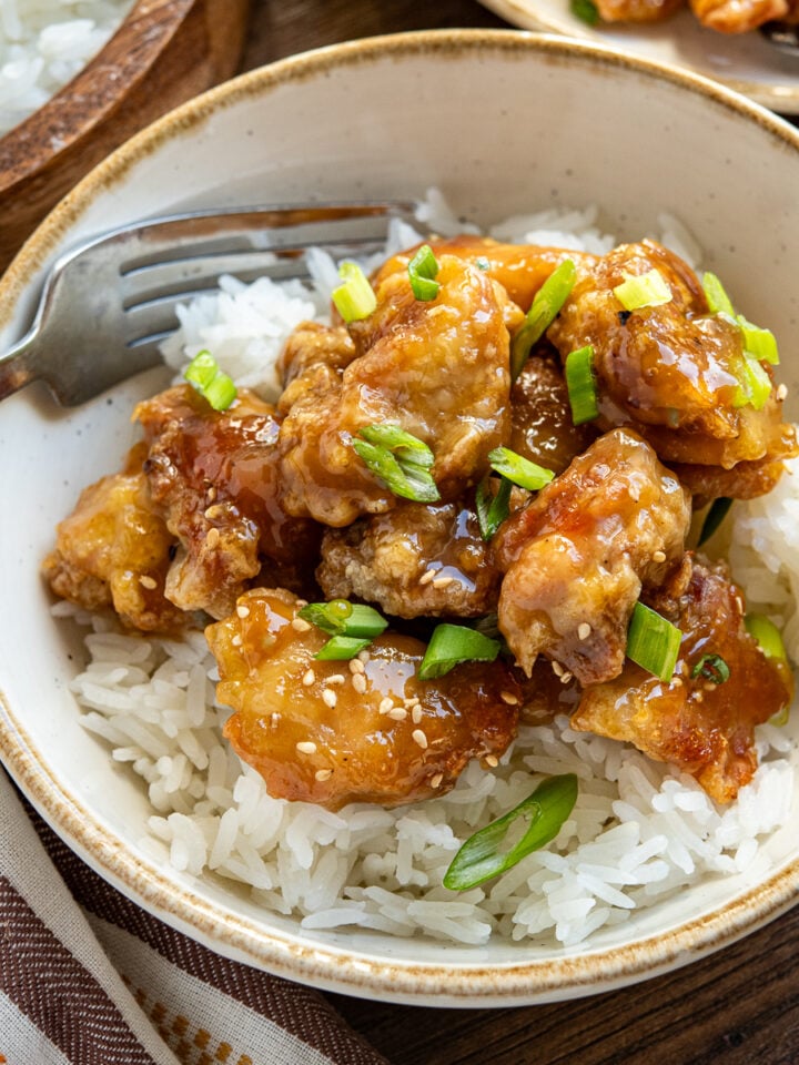 Chinese Lemon Chicken served in a white bowl with a beige rim, accompanied by fluffy white rice. A fork is elegantly placed on the left side of the bowl.