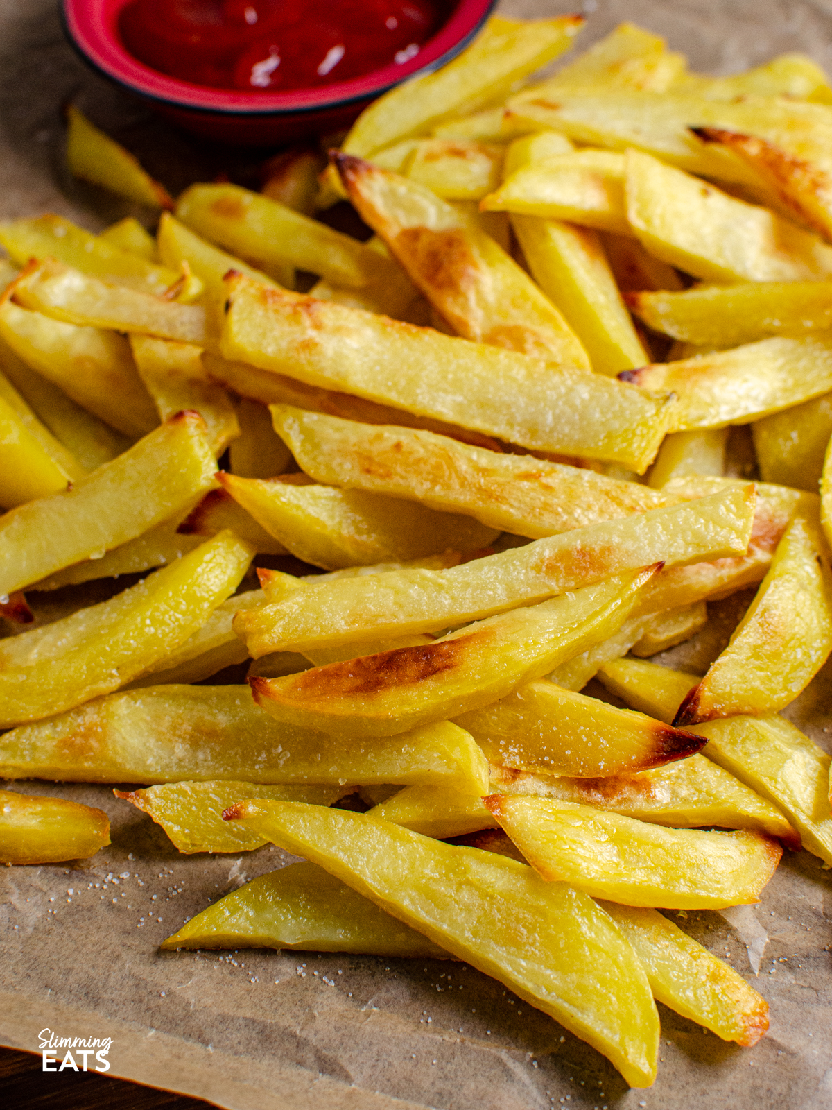 close up of Homemade Oven Baked Chip Shop Style Chips on parchment paper with a small red dish of ketchup