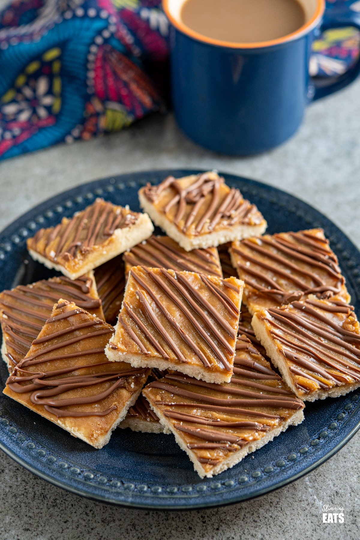 Millionaires Shortbread Squares on a dark blue navy plate with a blue mug of coffee