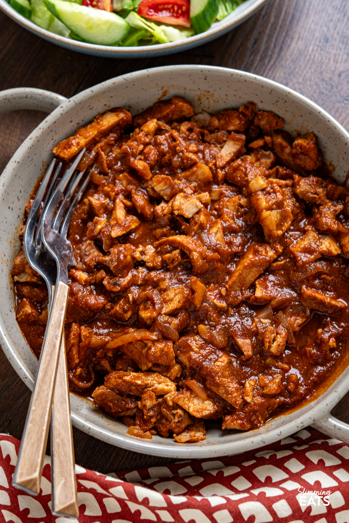 close up of stove top spicy bbq pork in a double handled brown grey ceramic dish, next to a bowl of salad 