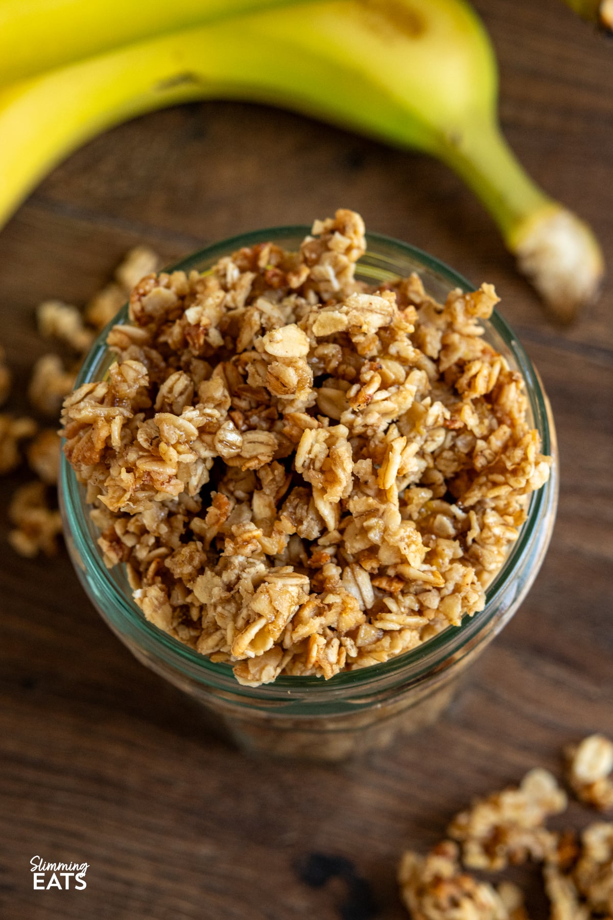 Indulge in the delightful flavours of this Banana Granola, a nutritious and tasty option that's perfect for kick-starting your day or as a scrumptious dessert. Crafted with wholesome oats, ripe bananas, and a touch of maple syrup, this granola is a fantastic low-calorie alternative that doesn't skimp on taste. close up of homemade banana granola in a jar on a wooden board surrounded by scattered granola and bananas
