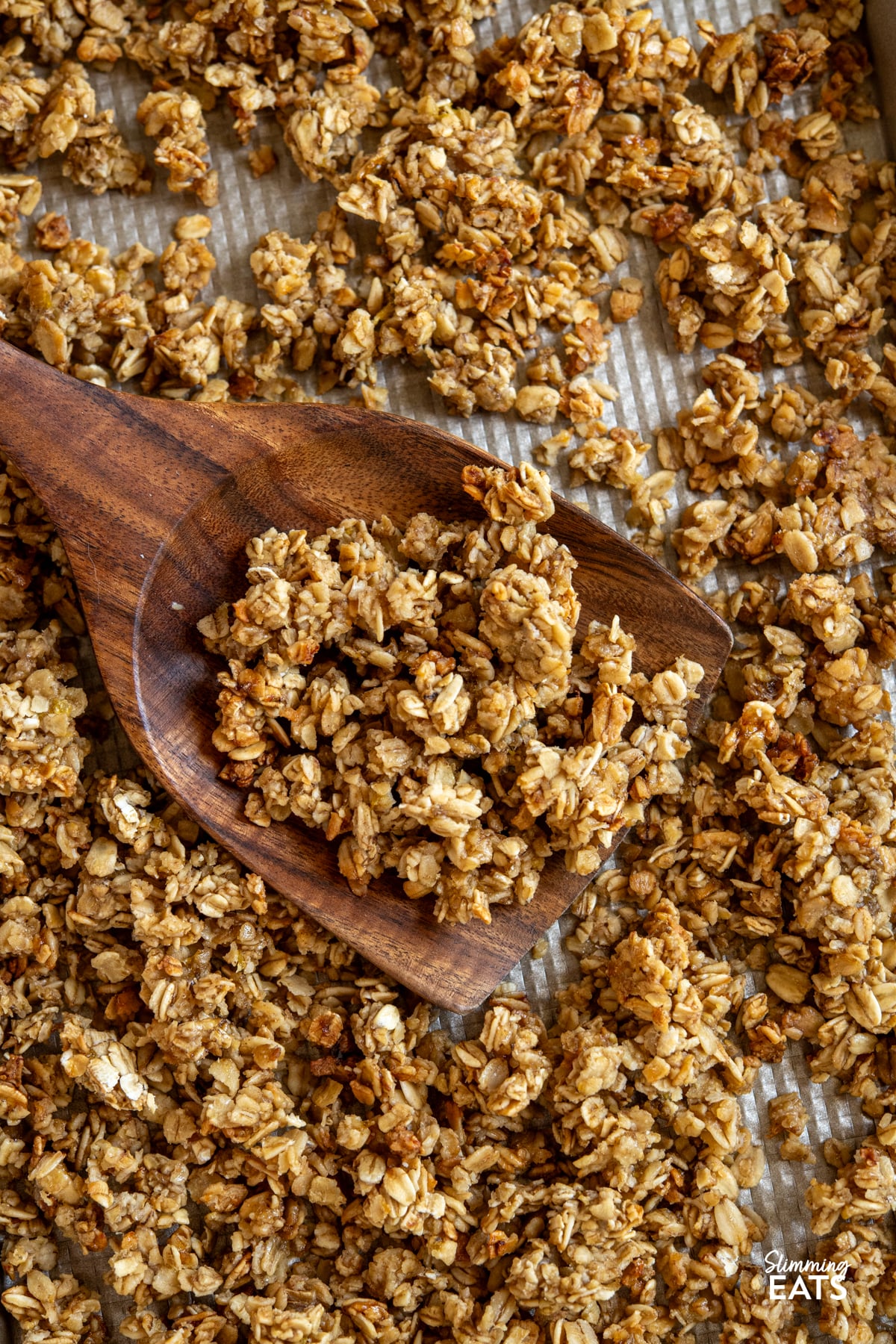 Indulge in the delightful flavours of this Banana Granola, a nutritious and tasty option that's perfect for kick-starting your day or as a scrumptious dessert. Crafted with wholesome oats, ripe bananas, and a touch of maple syrup, this granola is a fantastic low-calorie alternative that doesn't skimp on taste. wooden spatula scooping up homemade banana granola from a baking tray