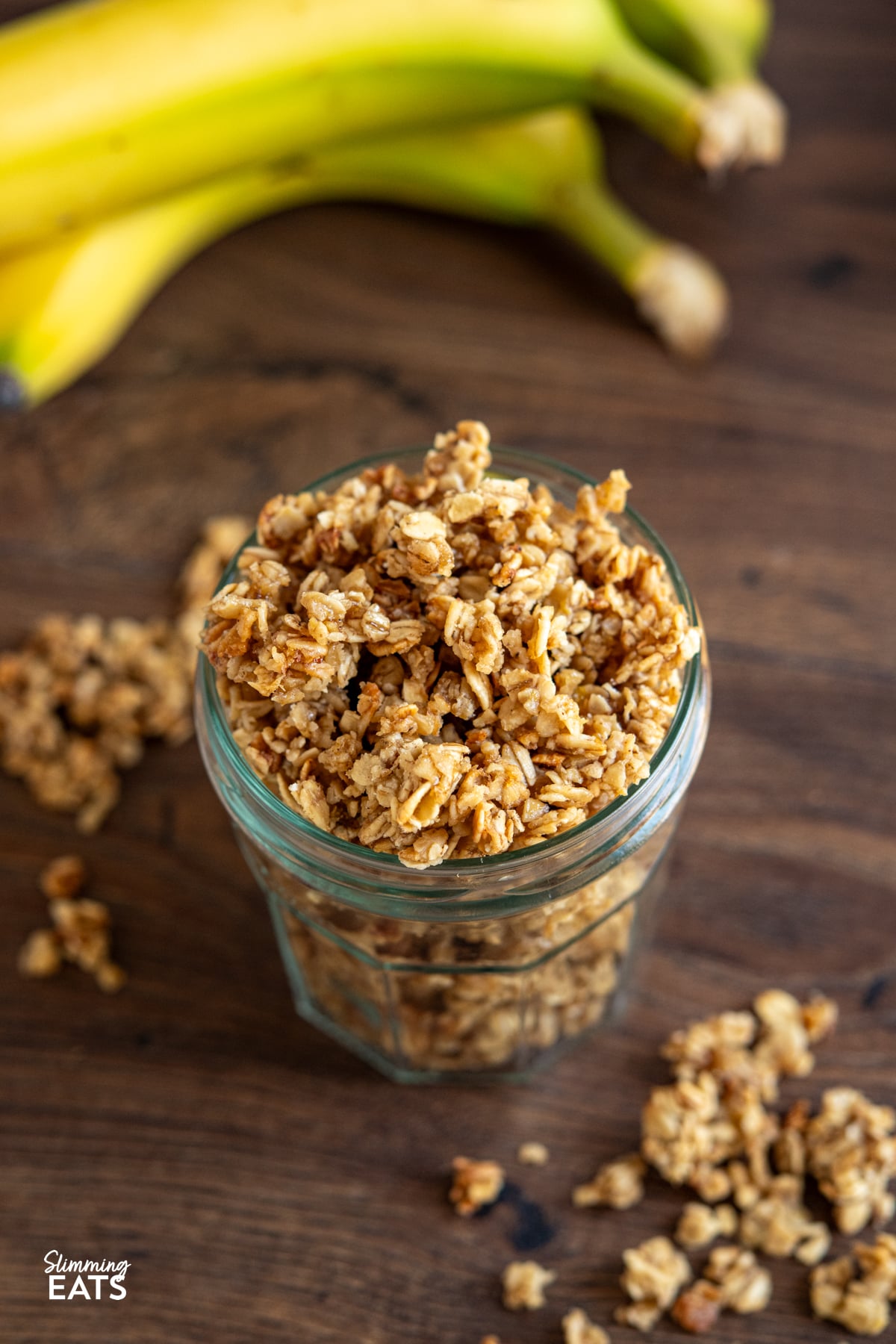 Indulge in the delightful flavours of this Banana Granola, a nutritious and tasty option that's perfect for kick-starting your day or as a scrumptious dessert. Crafted with wholesome oats, ripe bananas, and a touch of maple syrup, this granola is a fantastic low-calorie alternative that doesn't skimp on taste. homemade banana granola in a jar on a wooden board surrounded by scattered granola and bananas