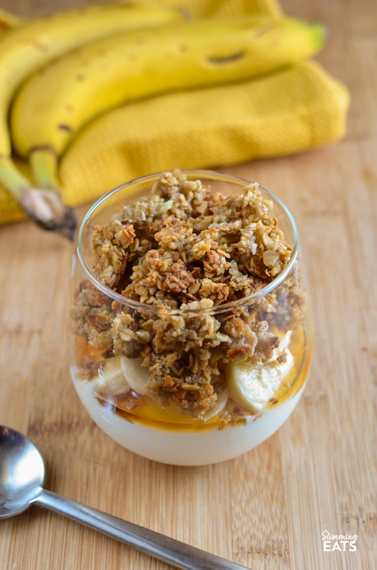 Banana Granola Parfaits - creamy yoghurt layered with homemade banana granola and fresh banana for an easy breakfast or sweet treat. The crunchy granola combined with the creamy yoghurt makes every spoonful so delicious and perfect any time of day. Banana Granola Yoghurt Parfait in a glass with bananas next to it on a yellow napkin.