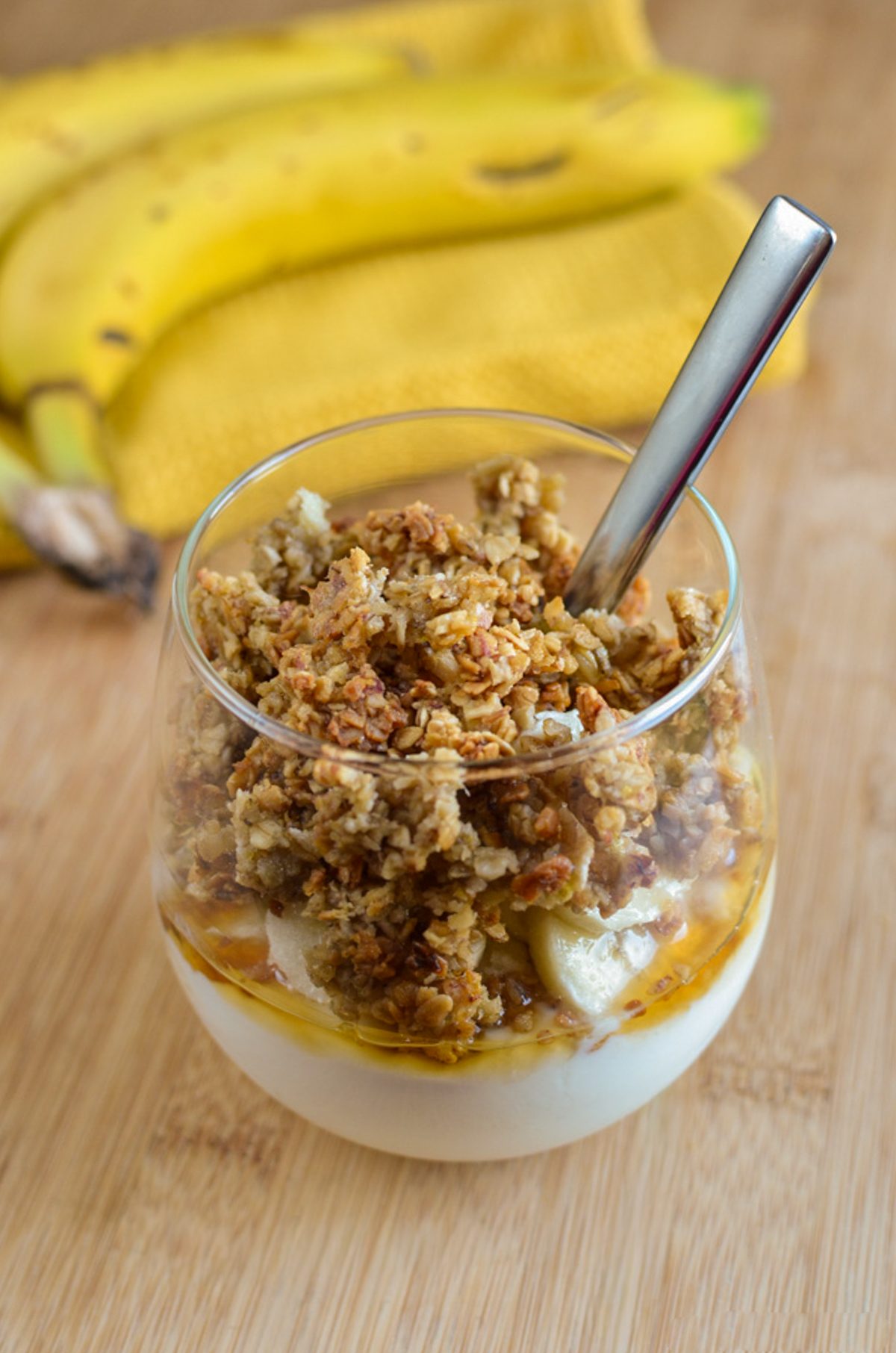 Banana Granola Parfaits - creamy yoghurt layered with homemade banana granola and fresh banana for an easy breakfast or sweet treat. The crunchy granola combined with the creamy yoghurt makes every spoonful so delicious and perfect any time of day. Banana Granola Yoghurt Parfait in a glass with spoon and bananas next to it on a yellow napkin.