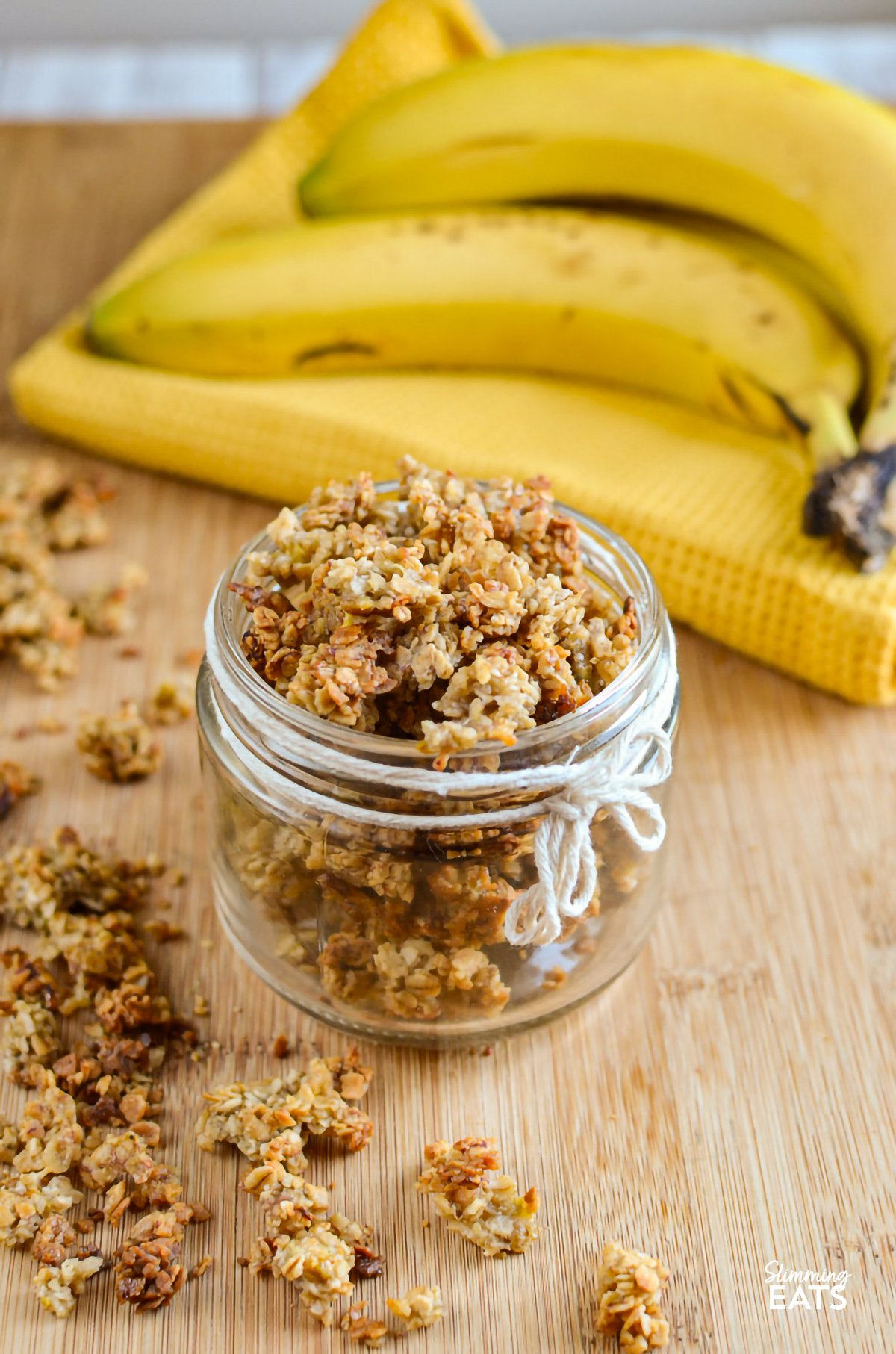 Banana granola beautifully presented in a small jar, decorated with a white rope thread tied around it, and whole bananas in the background for a natural, appetizing setting