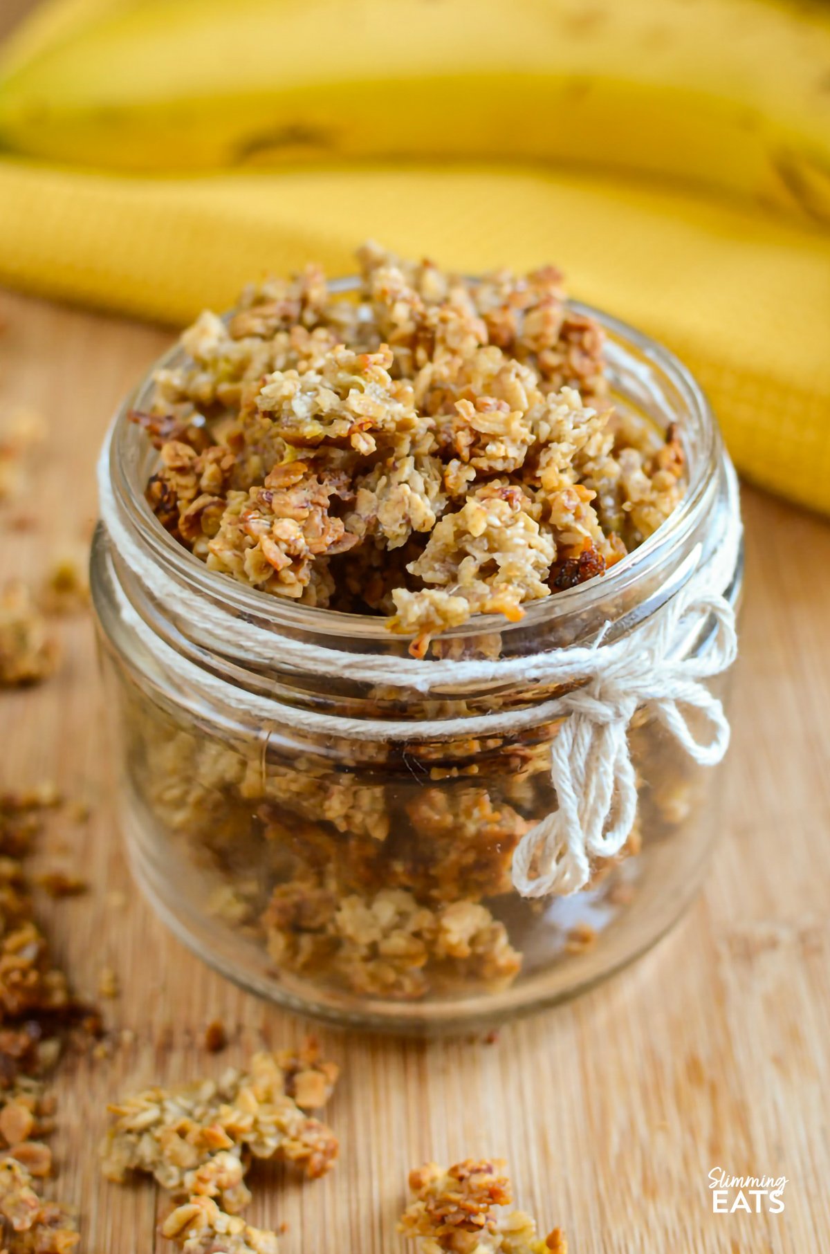 Banana granola beautifully presented in a small jar, decorated with a white rope thread tied around it, and whole bananas in the background for a natural, appetizing setting