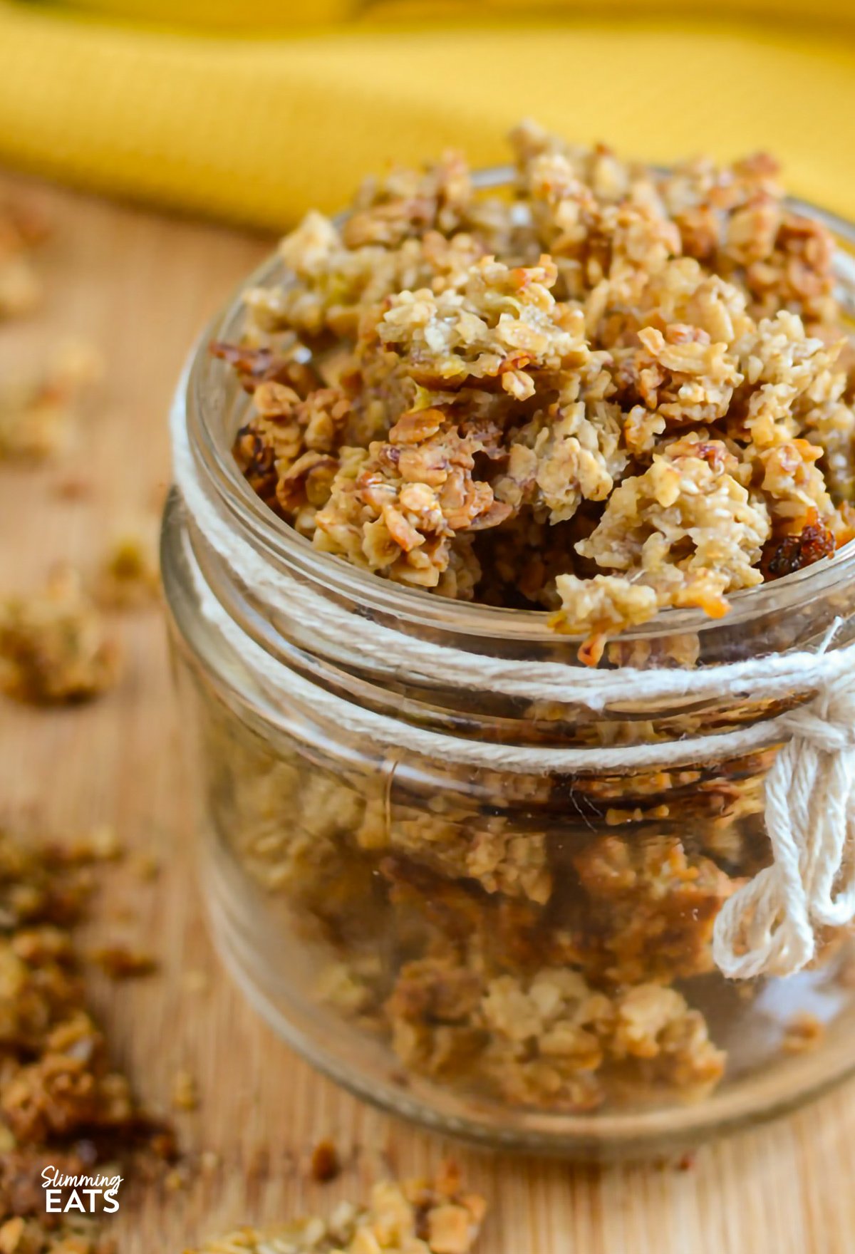 Banana granola beautifully presented in a small jar, decorated with a white rope thread tied around it, and whole bananas in the background for a natural, appetizing setting