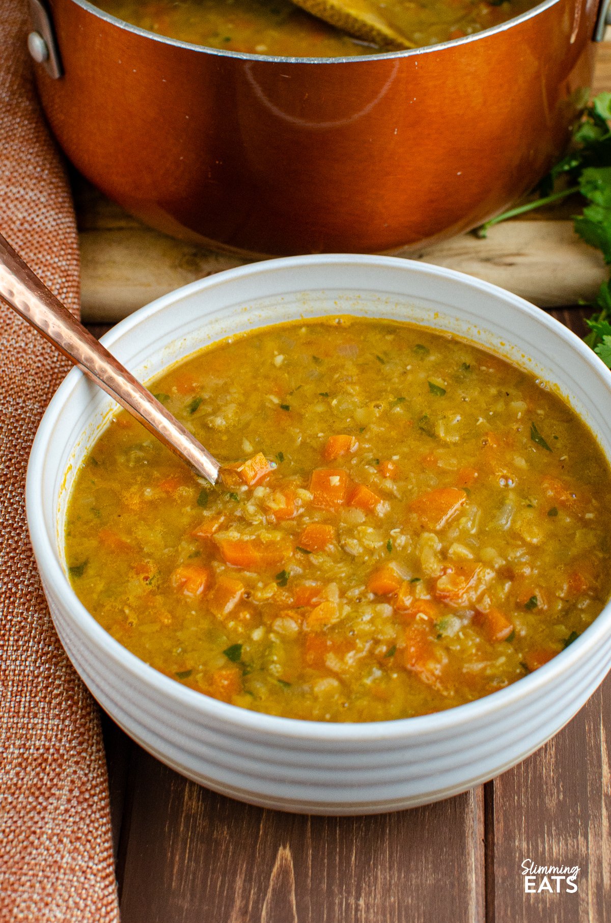 Spicy carrot and lentil soup served in a white bowl with a spoon, with the saucepan used for cooking visible in the background.