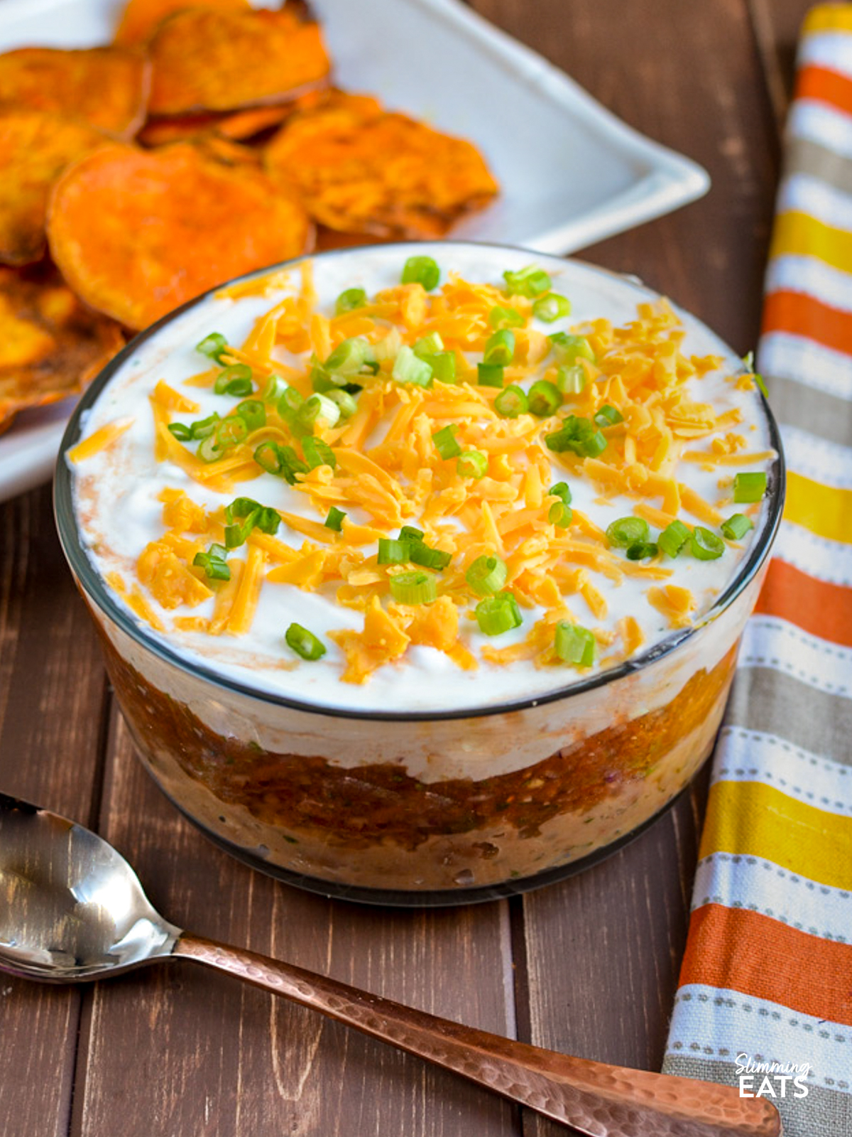 mexican layered dip in a bowl next to a yellow, orange, white and brown grey towel and a plate of homemade sweet potato chips